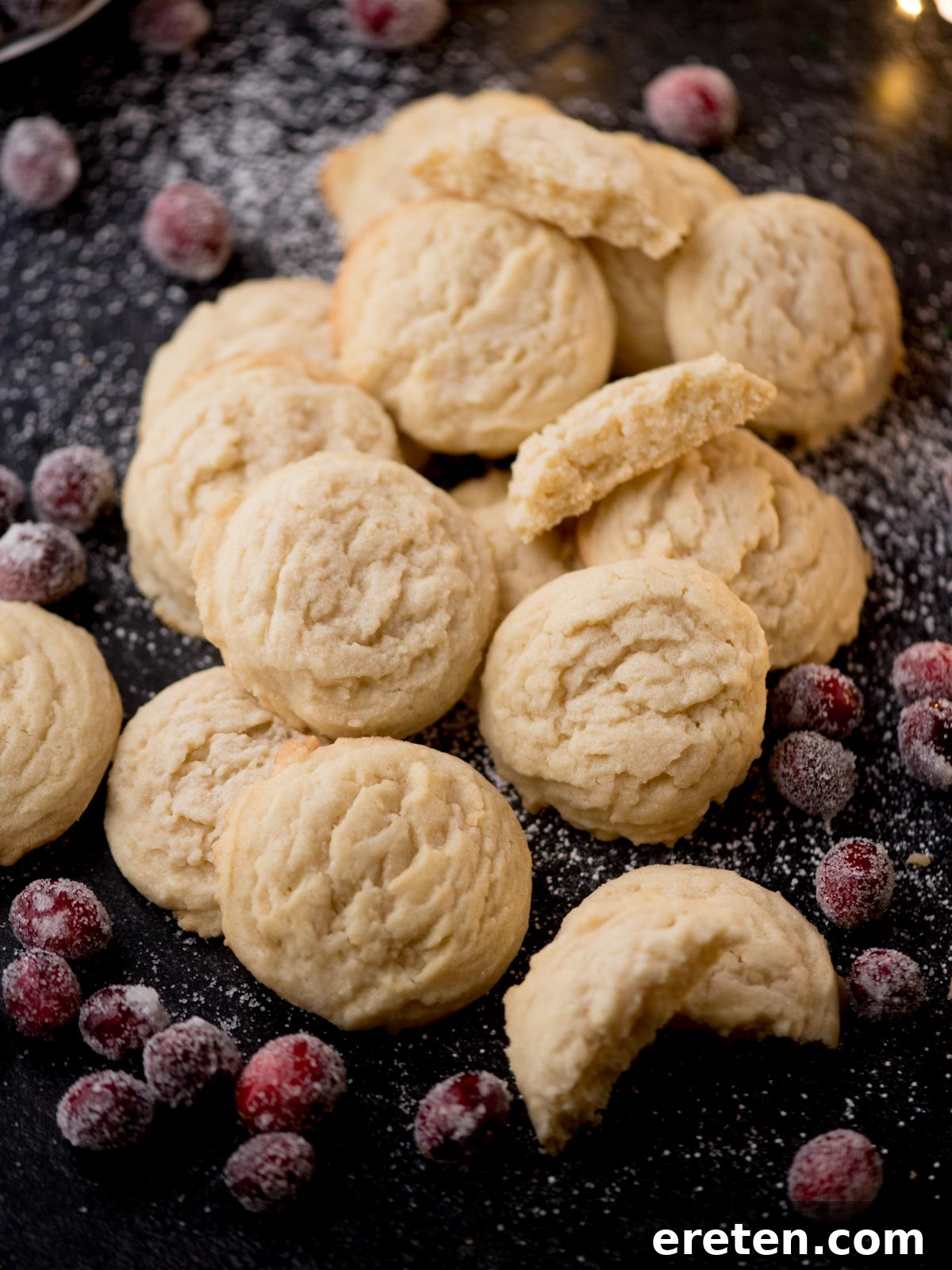 A pile of shortbread cookies is scattered on a dark surface, surrounded by sugared cranberries and a dusting of powdered sugar. Some cookies are whole, while a few are broken, revealing their crumbly texture reminiscent of the best Amish sugar cookies.