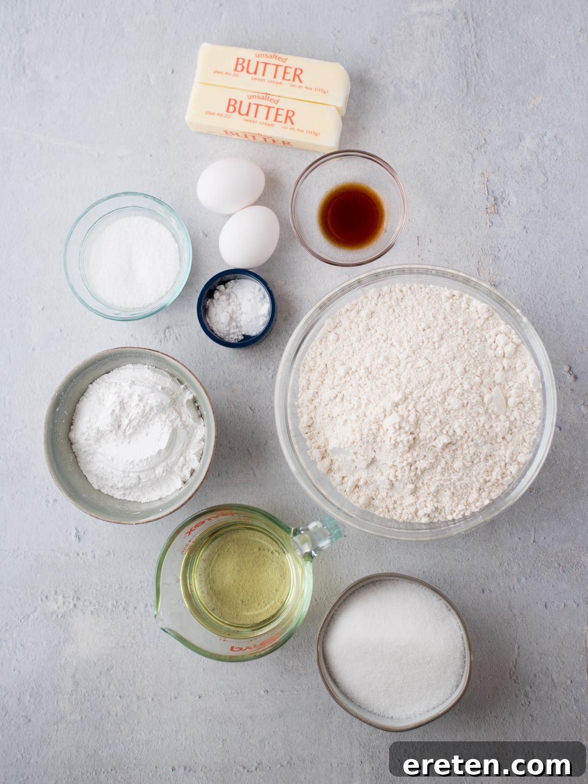 Ingredients for baking the best sugar cookies: two sticks of butter, two eggs, a bowl of flour, a bowl of sugar, a bowl of baking powder, a bowl of salt, a small bowl of vanilla extract, and a measuring cup of vegetable oil are all arranged on a light surface.