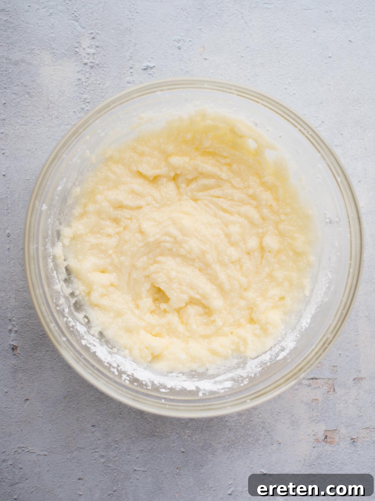 A clear glass bowl filled with a creamy, light yellow batter sits on a textured gray surface. The smooth mixture, with its few small lumps, promises the delectable taste of classic Amish sugar cookies in every bite.