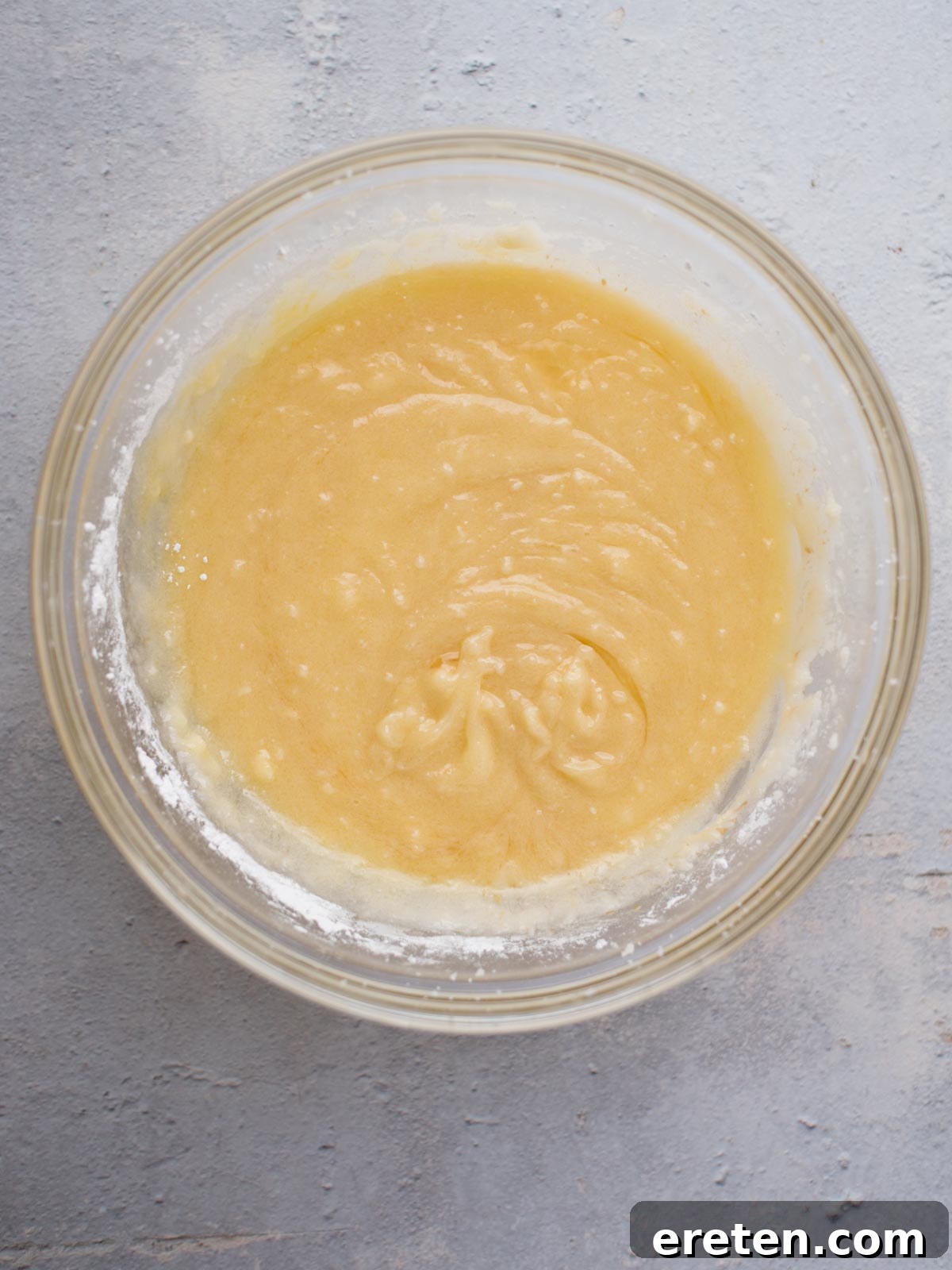 A glass bowl containing smooth, light yellow cake batter sits on a textured gray surface. The batter is slightly swirled in the center, resembling the rich mixture of Amish sugar cookies that have just been expertly combined.