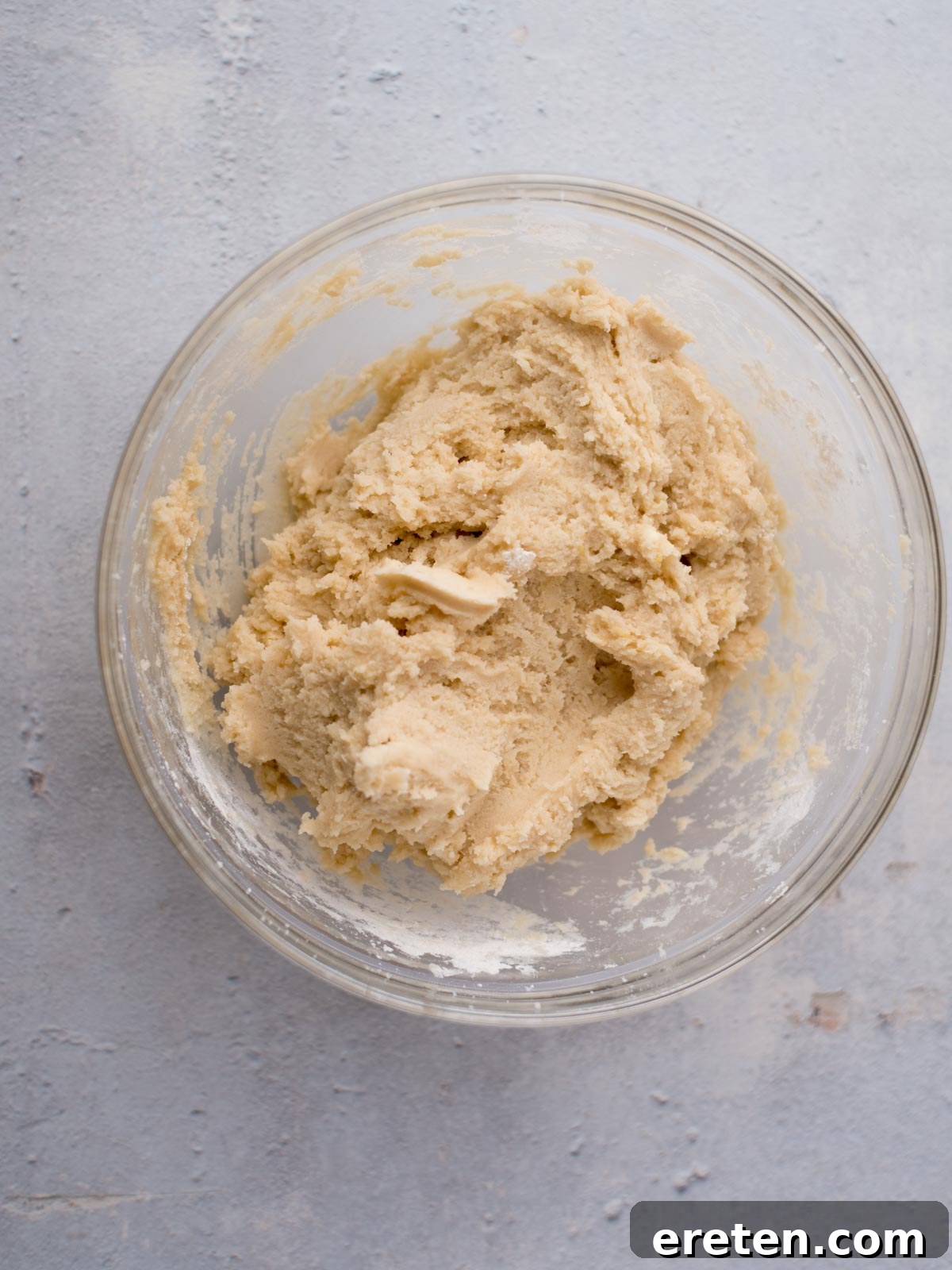 A glass bowl filled with unbaked Amish sugar cookie dough sits on a textured light gray surface. The dough appears creamy and thick, mixed to a smooth consistency that promises the best sugar cookies.