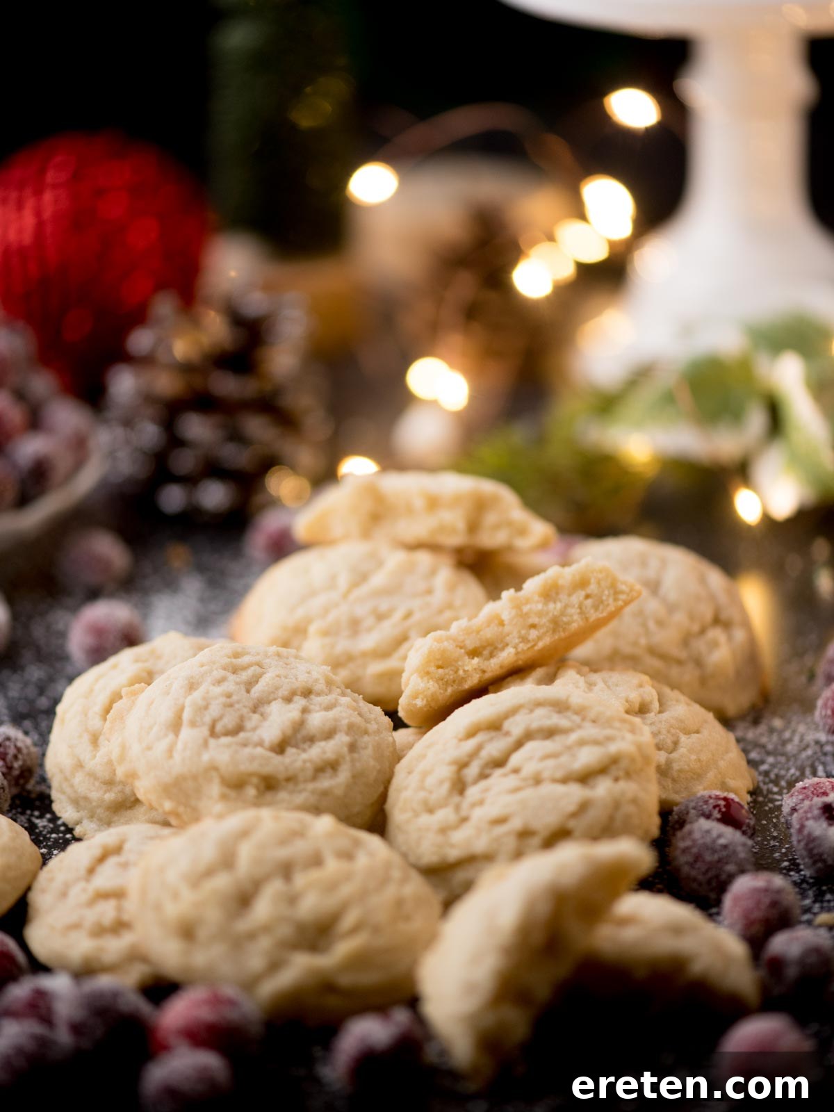 A pile of the best sugar cookies is surrounded by sugared cranberries on a festive table. The background features blurred twinkling lights and a red ornament, creating a cozy holiday atmosphere.