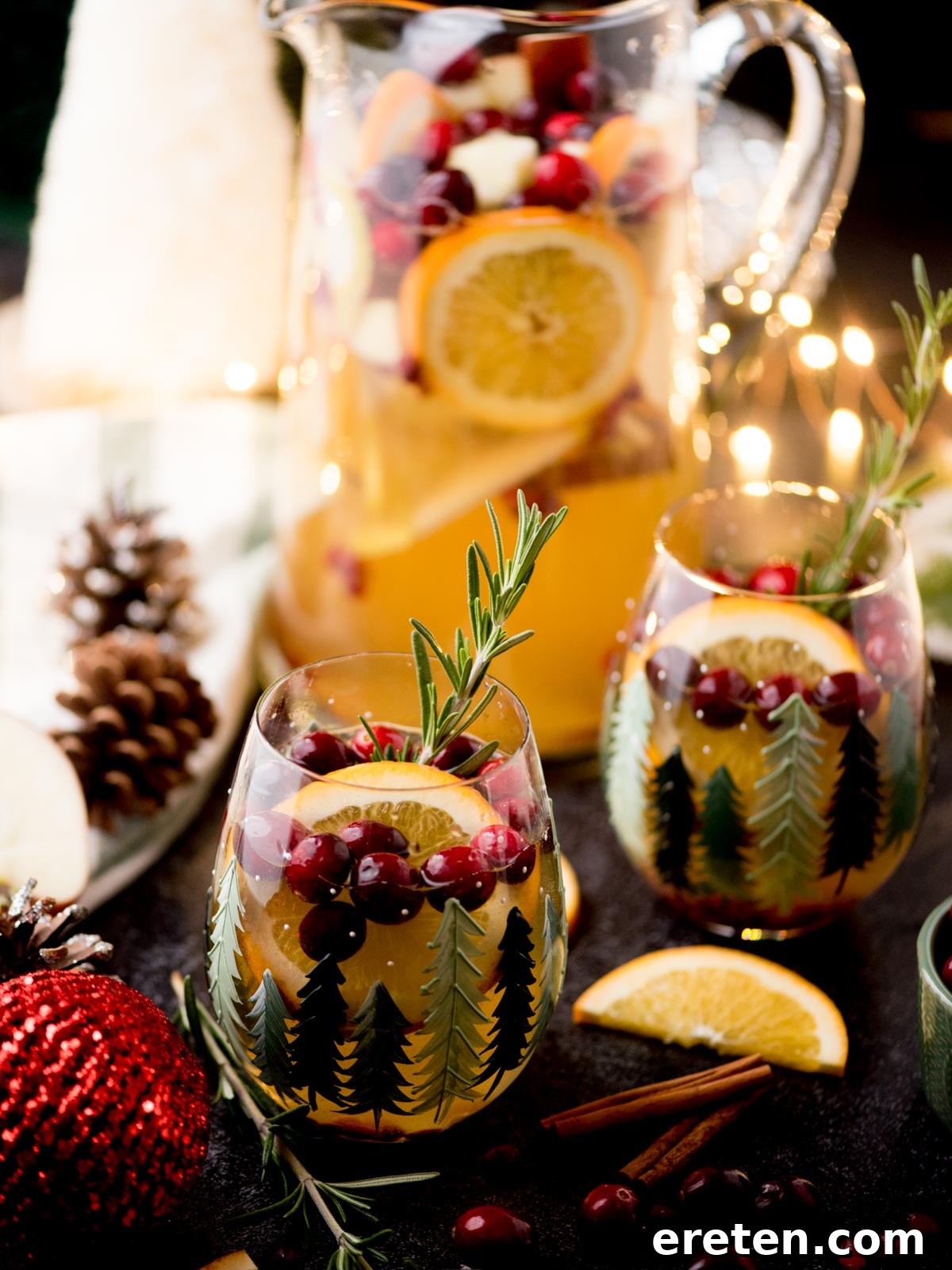 A festive White Christmas Sangria served in pine-themed glasses, beautifully garnished with ruby-red cranberries and aromatic rosemary sprigs. In the background, a large pitcher overflows with sliced apples, oranges, and more cranberries, surrounded by charming holiday decorations such as pinecones, cinnamon sticks, and dried orange slices.