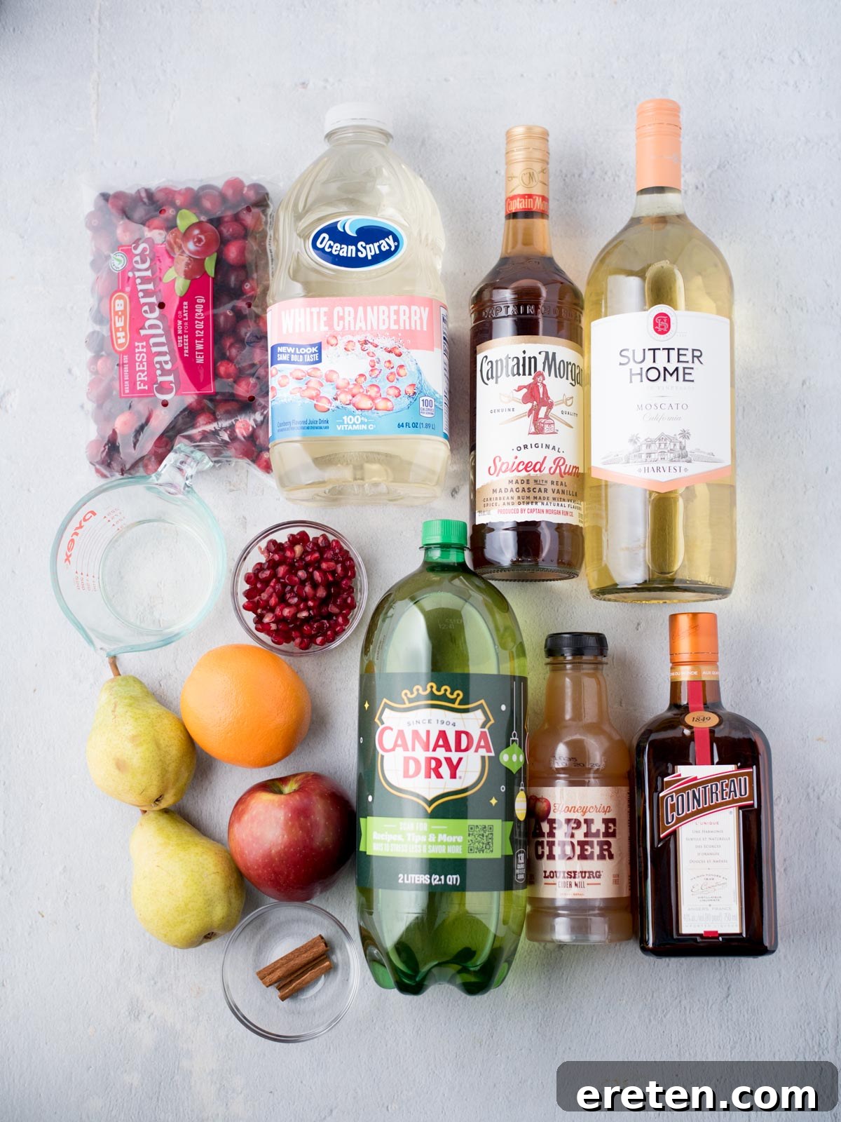 An overhead view of a countertop laden with all the ingredients for White Christmas Sangria. Visible are bottles of white cranberry juice, Captain Morgan spiced rum, Sutter Home Moscato, and Canada Dry ginger ale. Fresh produce includes a bag of bright red cranberries, a ripe orange, and green pears. Also present are a bottle of Cointreau, apple cider, a container of pomegranate seeds, and several cinnamon sticks, all ready for preparation.