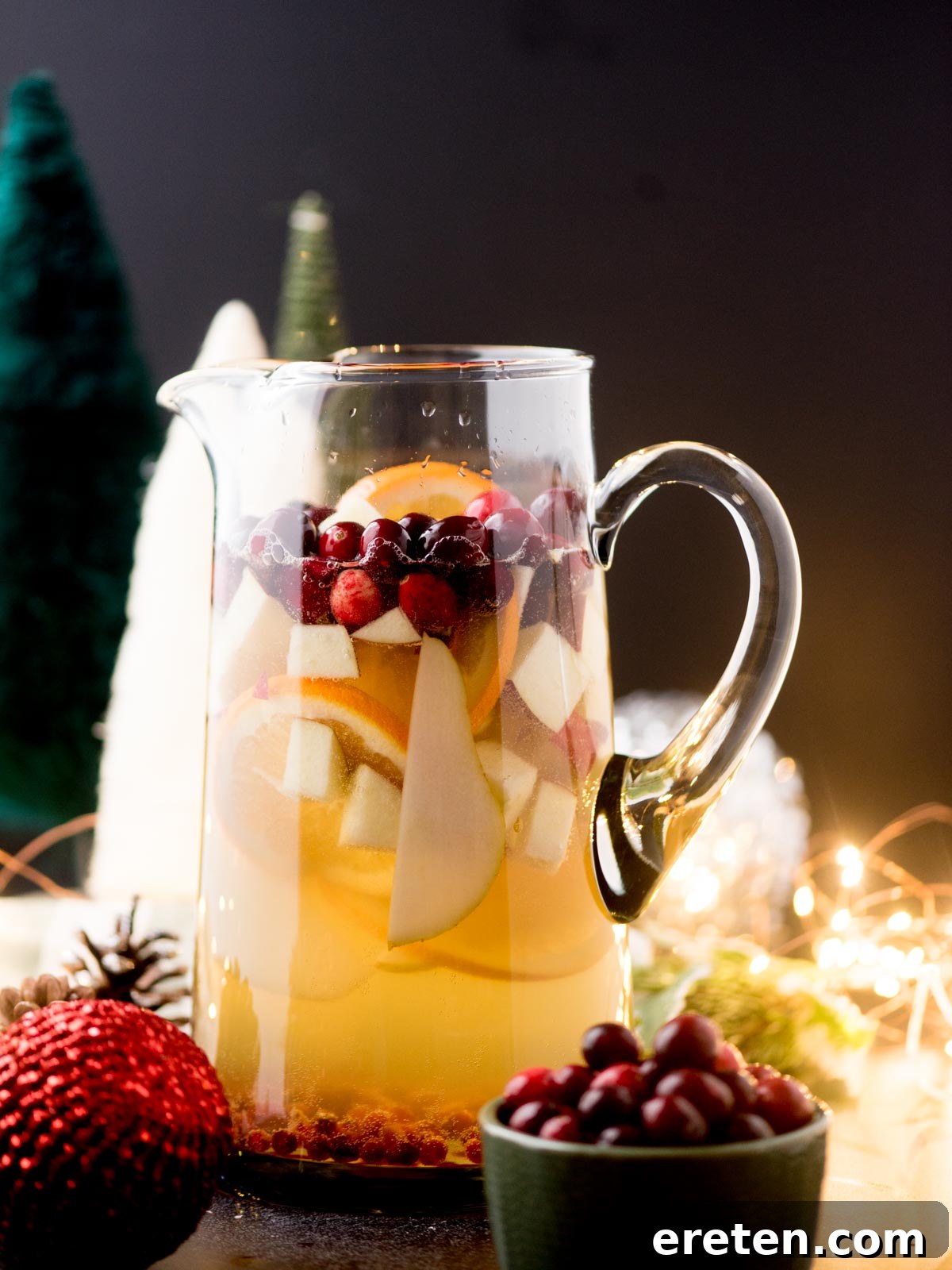 A beautifully arranged glass pitcher filled with festive Christmas Sangria, featuring visible slices of apples, oranges, and a scattering of bright red cranberries. The pitcher is surrounded by charming holiday decorations, including natural pine cones, shiny Christmas ornaments, and delicate, soft-lit string lights, creating a warm and inviting atmosphere.