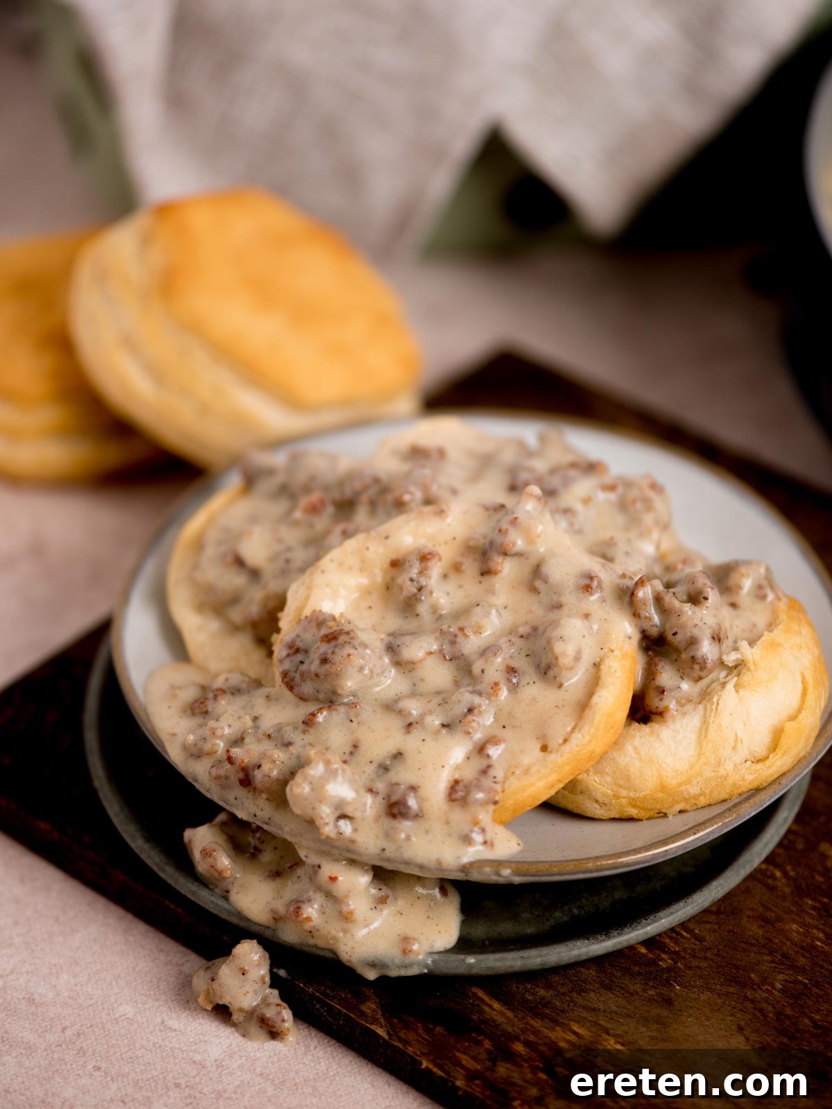 A plate of freshly baked biscuits topped with creamy sausage gravy. Additional biscuits are seen in the background on a wooden surface. The scene suggests a hearty, homemade breakfast setting.