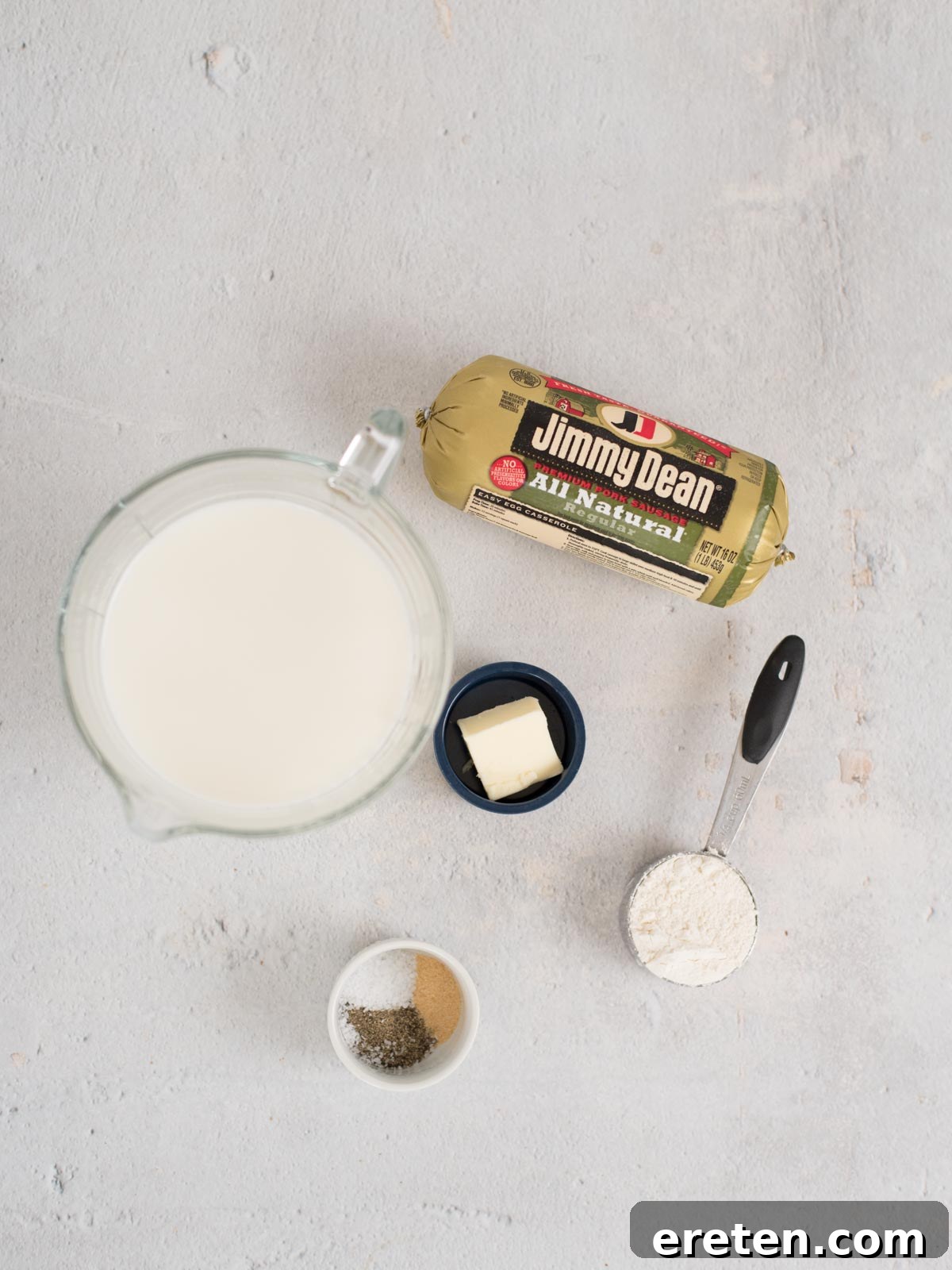 A top view of breakfast gravy ingredients on a light surface: a package of Jimmy Dean sausage, a pitcher of milk, a small bowl with butter, a measuring cup with flour, and a small dish containing salt and pepper.