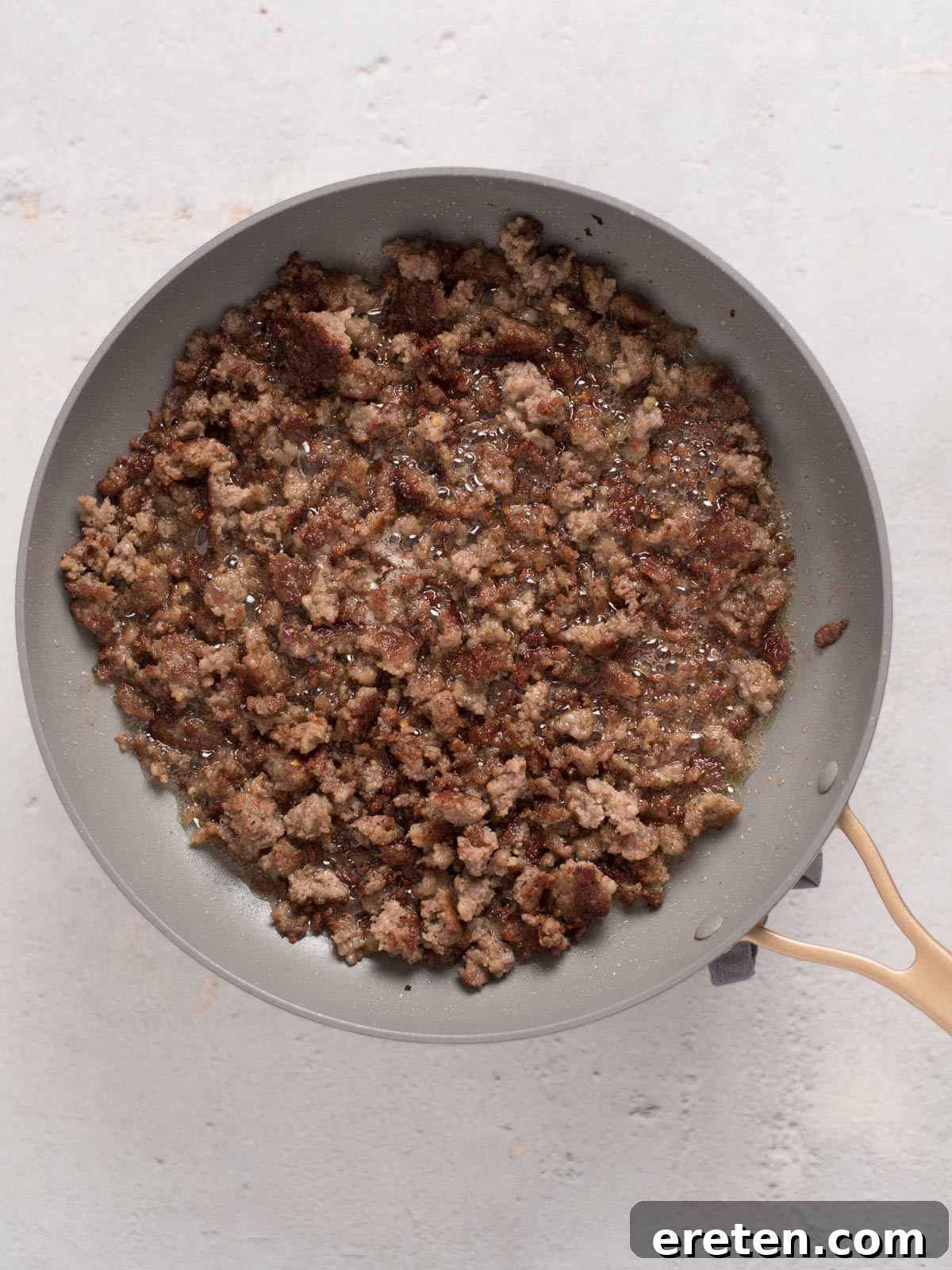 A pan containing browned and crumbled ground meat is placed on a light surface. The meat is cooked evenly, with a slightly crispy texture. The pan has a light gray interior and a wooden handle.