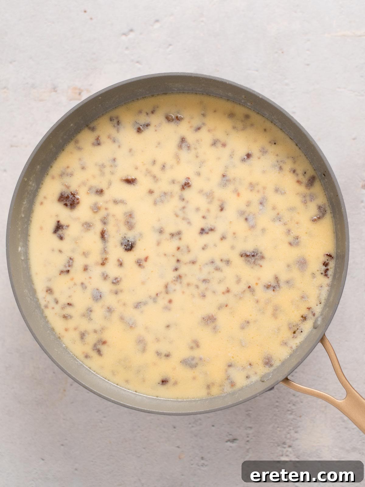 A pan filled with a creamy yellow mixture speckled with chunks of ground sausage. The mixture has a smooth, slightly thick texture and is set against a light gray background.