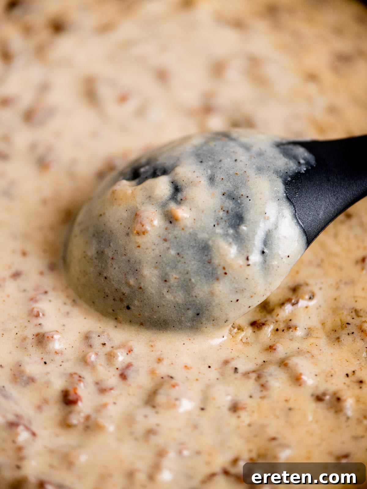 Close-up of a creamy gravy with ground sausage bits, being stirred by a black spoon. The gravy has a speckled appearance, indicating the presence of pepper or spices.