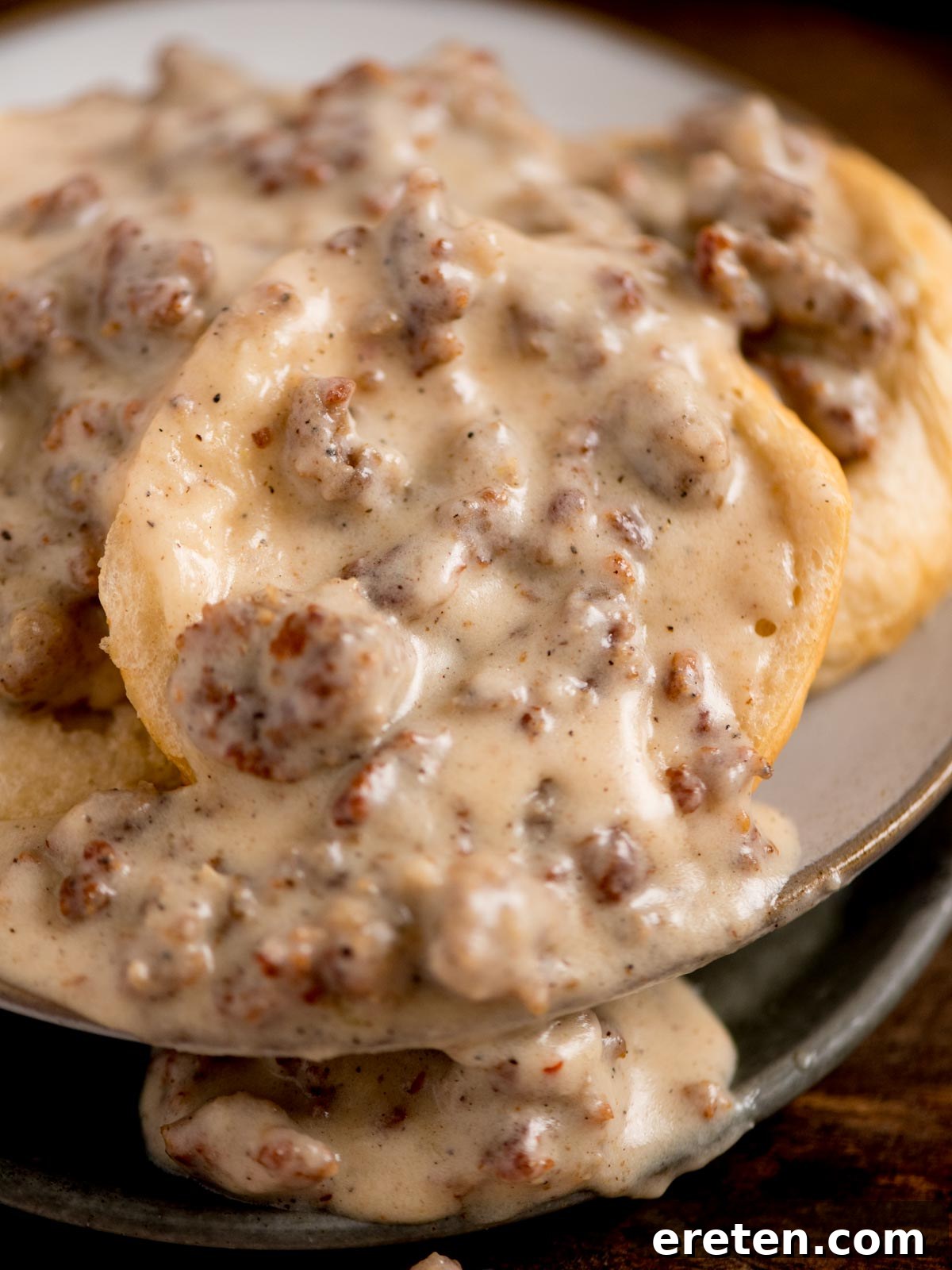 A close-up of a dish featuring biscuits topped with creamy sausage gravy. The gravy is thick and speckled with pieces of crumbled sausage, covering the golden-brown biscuits on a plate.