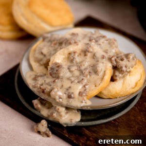 A plate of biscuits covered with creamy sausage gravy. The biscuits are fluffy and golden, with pieces of cooked sausage visible in the thick, peppered gravy. Another biscuit is in the background on the wooden surface.