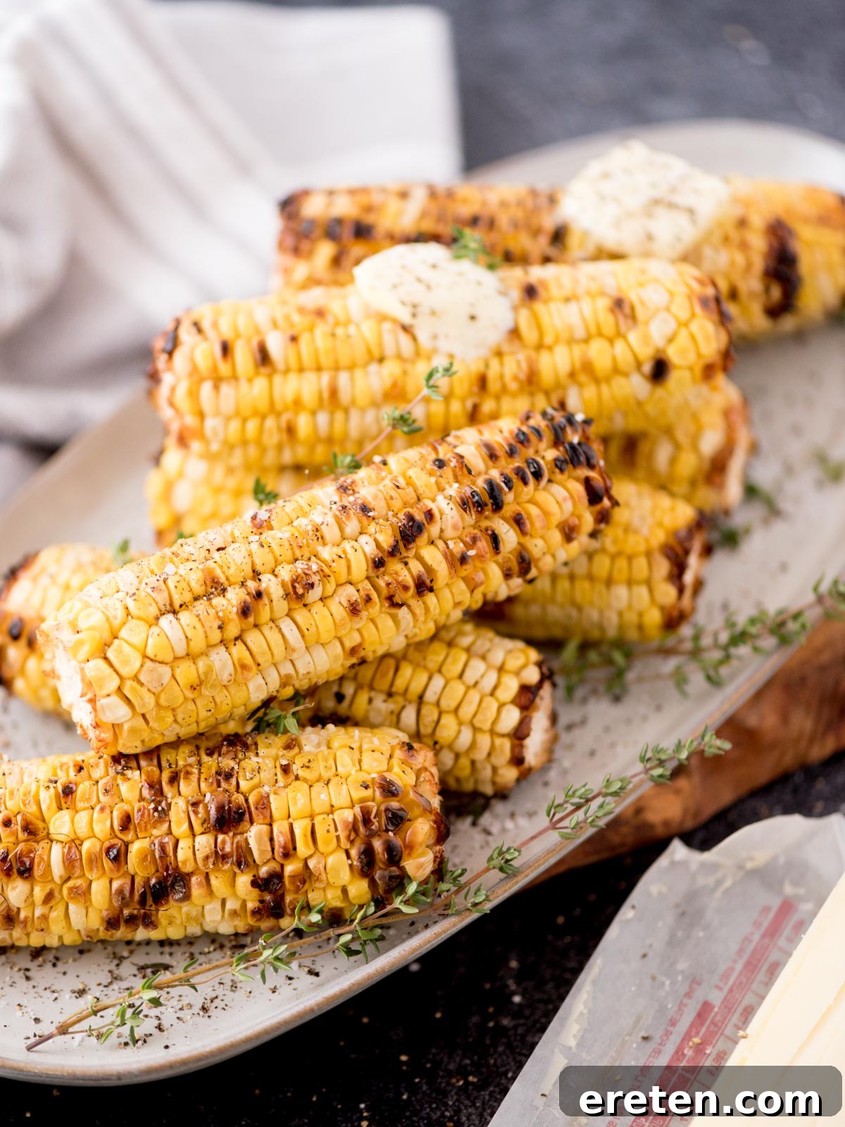 Grilled corn on the cob stacked on a plate, topped with pats of butter and garnished with fresh thyme. A white cloth napkin is in the background.