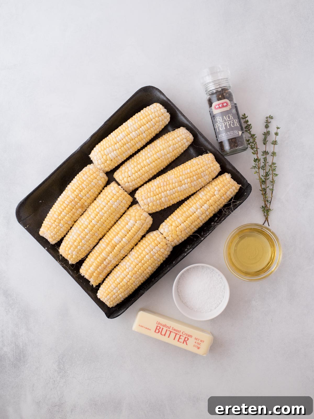 Nine ears of corn on a black tray, with a stick of butter, a bowl of salt, a small bowl of oil, a sprig of thyme, and a bottle of black pepper arranged on a white surface.