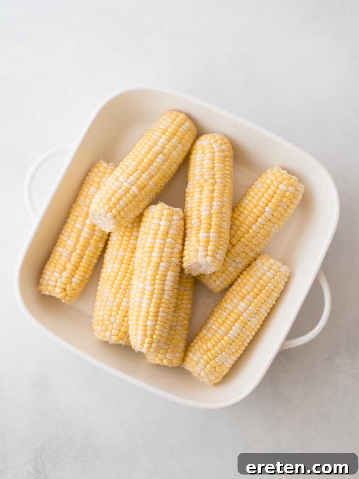 A white dish filled with several uncooked, halved ears of corn, neatly arranged on a light-colored surface.