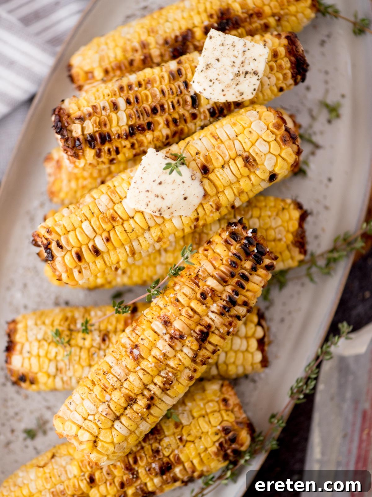 Grilled corn on the cob stacked on a plate, topped with melting butter, fresh thyme, and a sprinkle of black pepper.