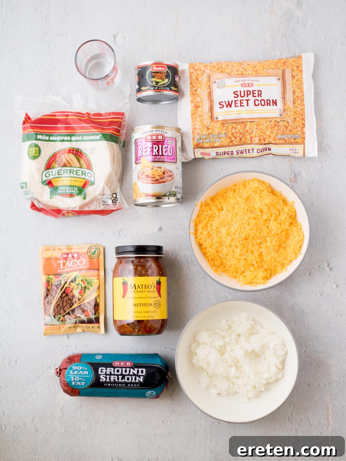Top view of ingredients on a white surface: corn tortillas, refried beans, taco seasoning mix, salsa, ground sirloin, diced onions, shredded cheese, super sweet corn, and a small glass of water.
