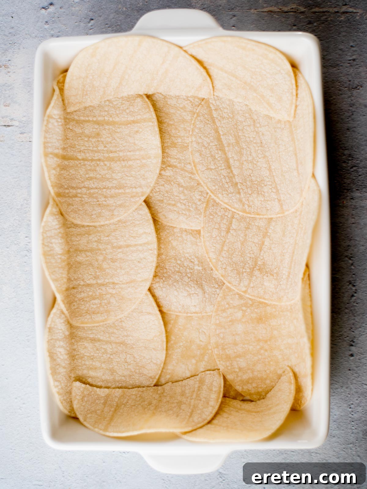 A rectangular baking dish filled with overlapping layers of round corn tortillas, placed on a textured gray surface.