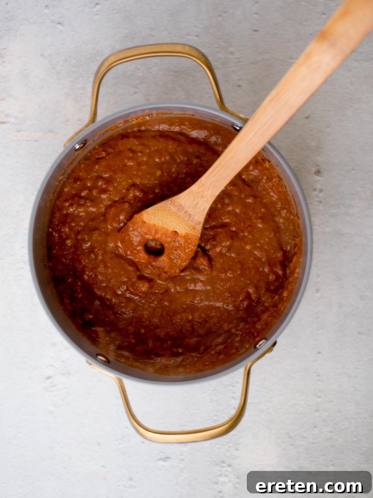 A pot of thick, brown chili with a wooden spoon stirring it, viewed from above. The pot has gold handles and rests on a grey surface. The chili appears rich and textured.