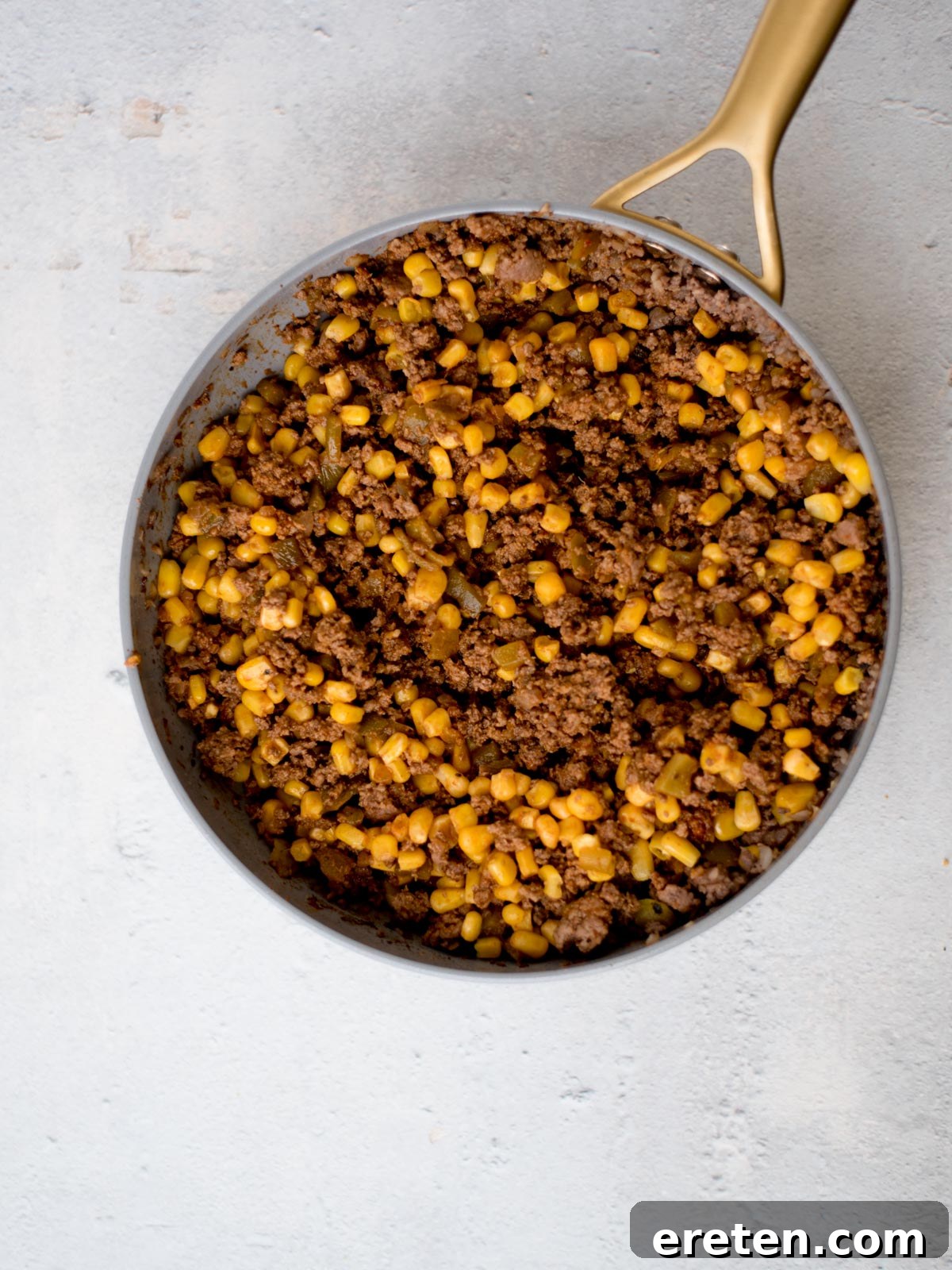 A skillet filled with cooked ground beef and corn kernels on a light gray surface. The ingredients are mixed together, giving a brown and yellow appearance. The skillet has a gold-colored handle.