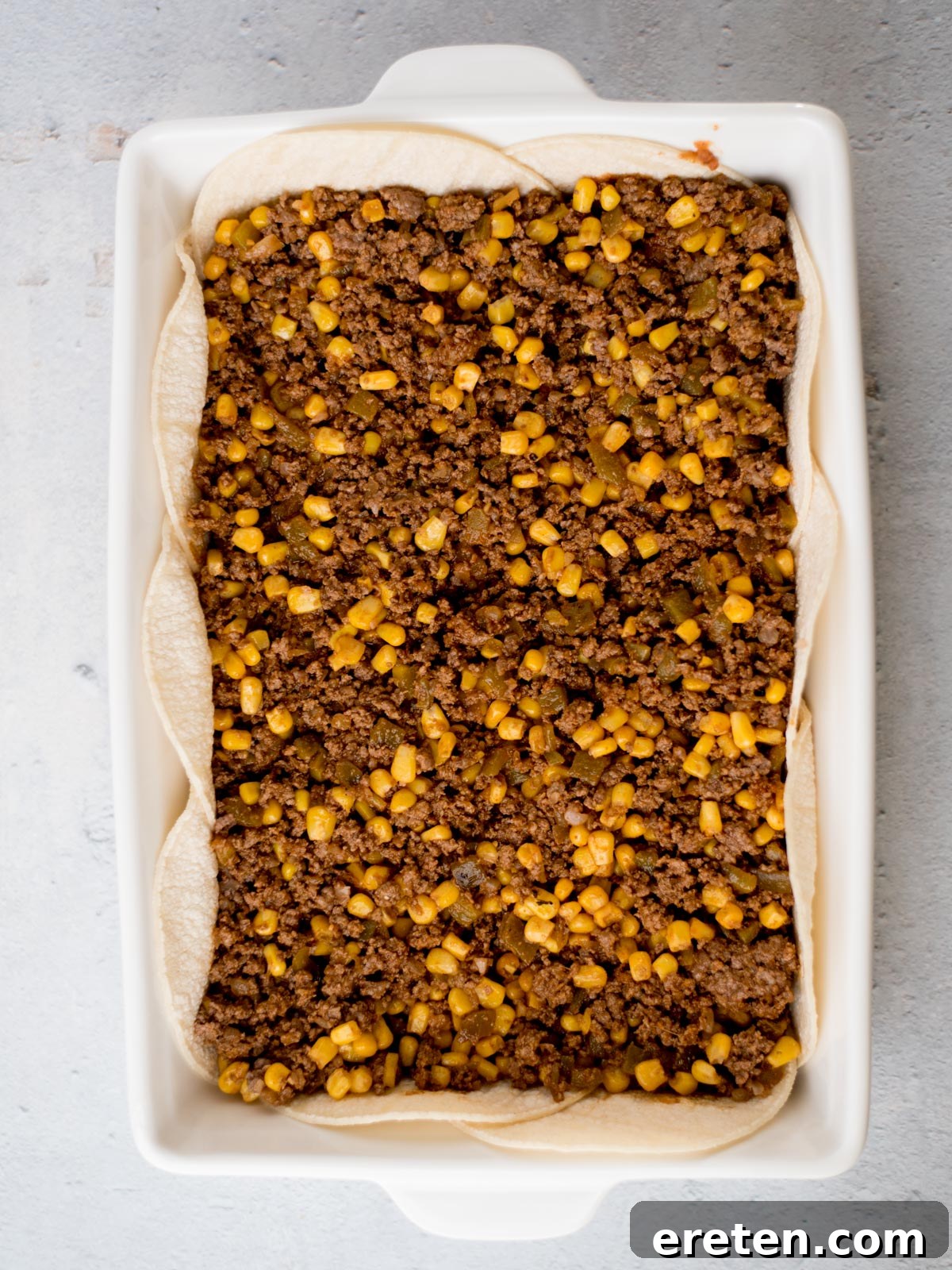 A rectangular baking dish filled with layered tortillas topped with seasoned ground beef and corn. The dish is ready for the oven, resting on a light gray surface.