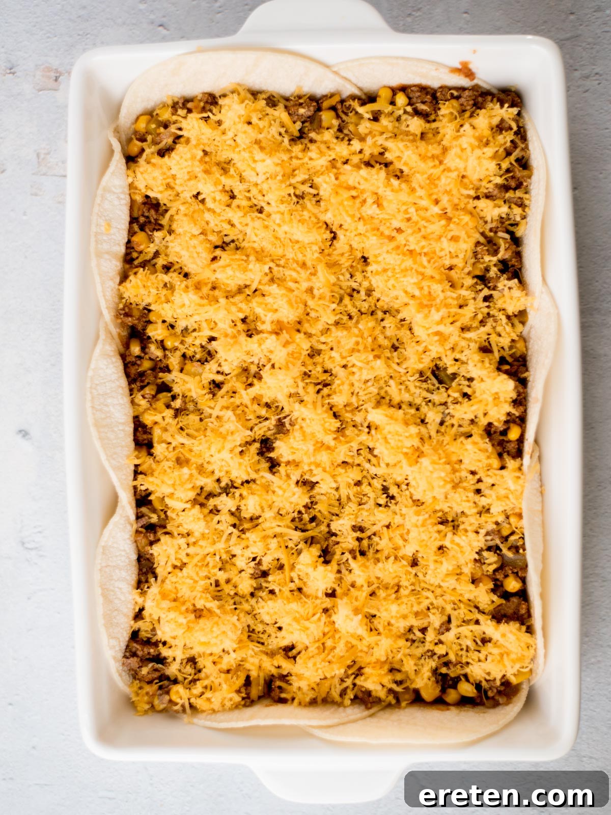 A rectangular baking dish filled with layers of tortillas, ground beef, corn, and melted cheese, creating a casserole. The dish is placed on a light gray surface.