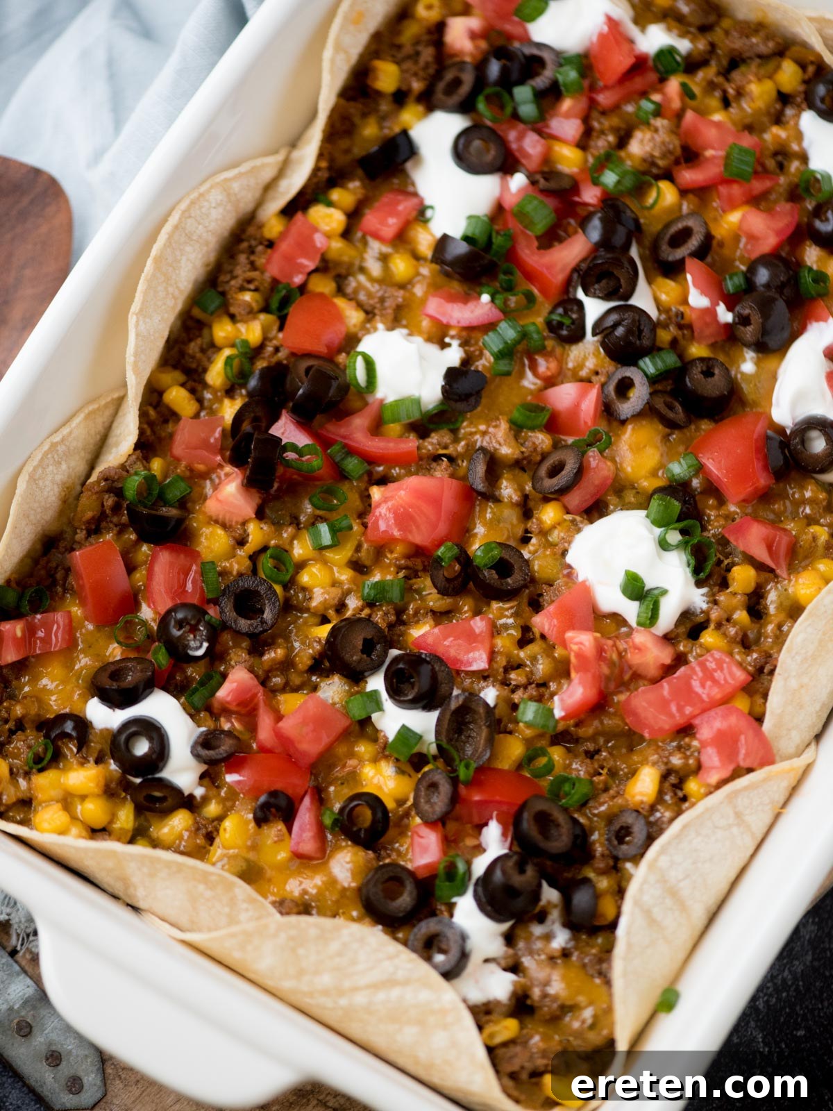 A casserole dish filled with a layered taco bake, featuring tortillas, ground meat, corn, melted cheese, diced tomatoes, black olives, sour cream dollops, and green onions. The edges are partially folded over the filling.