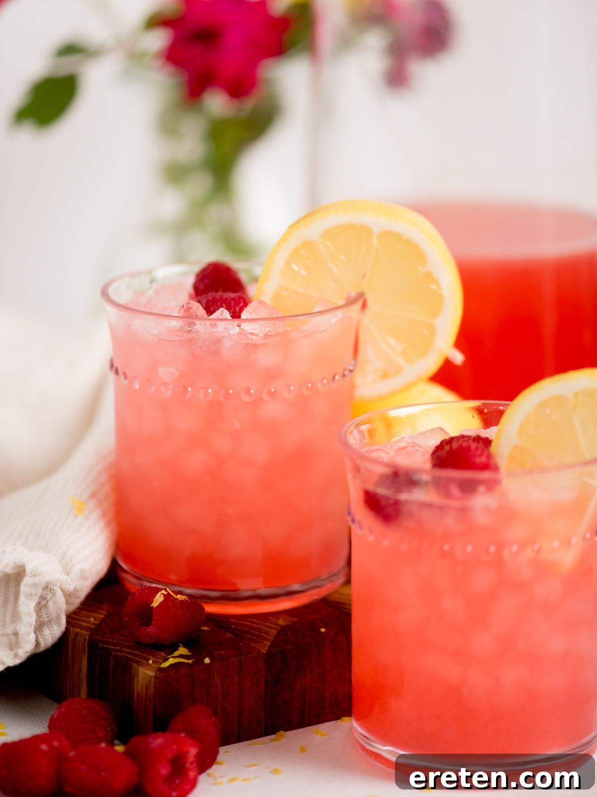 Two glasses of vibrant pink homemade raspberry lemonade, brimming with ice, are elegantly garnished with fresh lemon slices and whole raspberries. They rest on a rustic wooden board, flanked by scattered fresh raspberries and a soft white cloth, with a background of subtly blurred greenery and flowers, suggesting a serene summer setting.