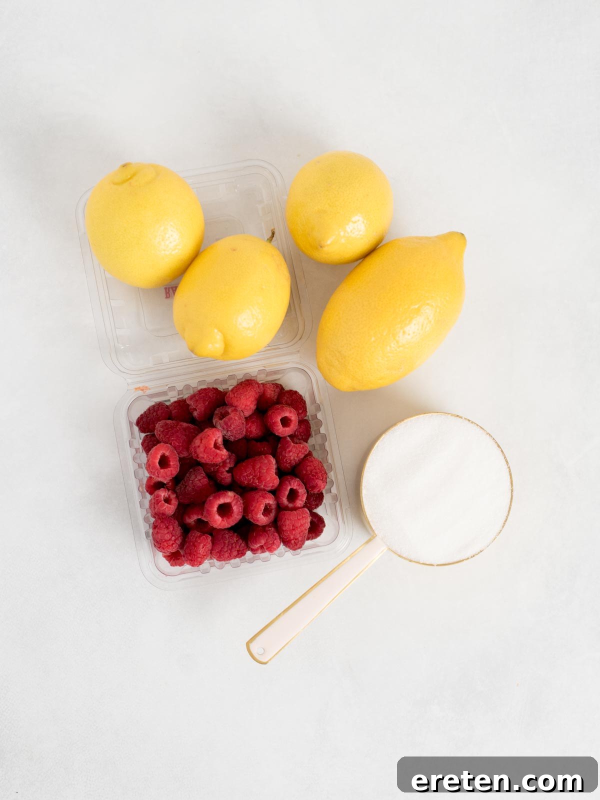 A bright and inviting flat lay of essential ingredients for raspberry lemonade: four glistening yellow lemons, a clear plastic container brimming with fresh, ripe red raspberries, and a glass measuring cup filled with sparkling granulated sugar. All are neatly arranged on a pristine white surface, highlighting their freshness and quality.