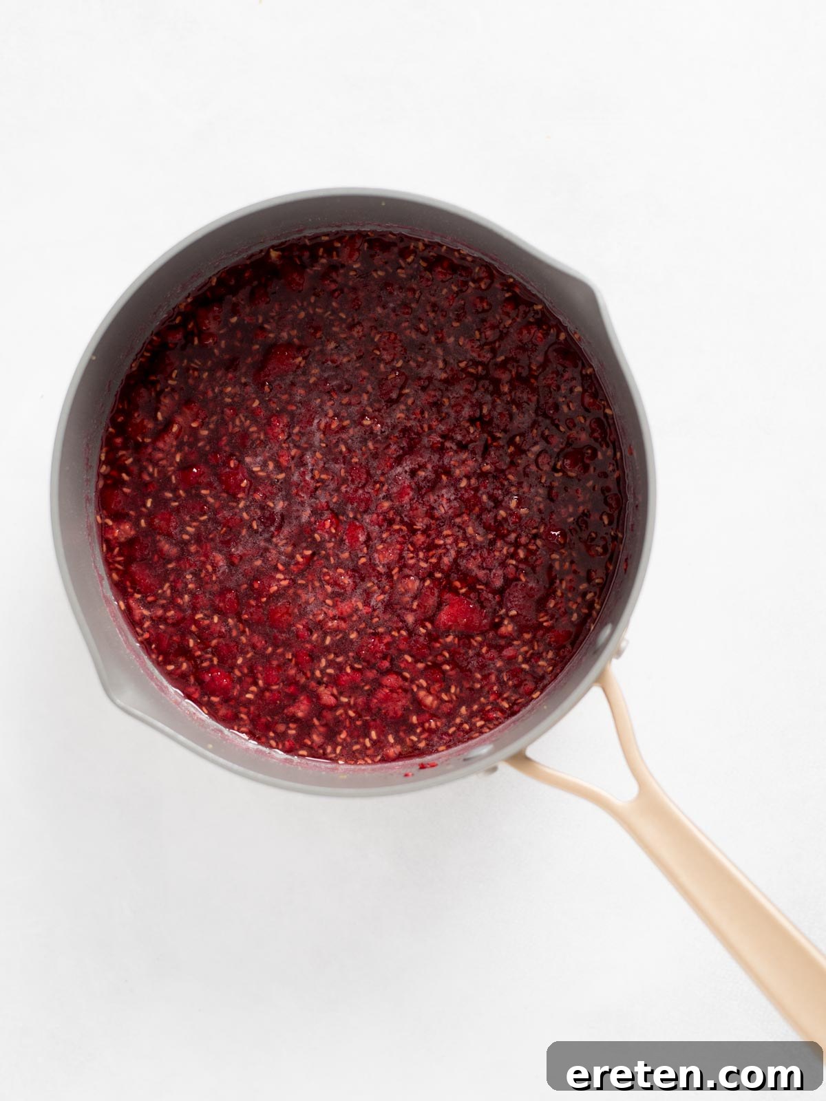 A gray saucepan with a beige handle, brimming with a rich, simmering raspberry sauce or jam, elegantly positioned on a pristine white surface. The mixture exhibits a thick, glossy texture, hinting at the intense fruit flavor within.