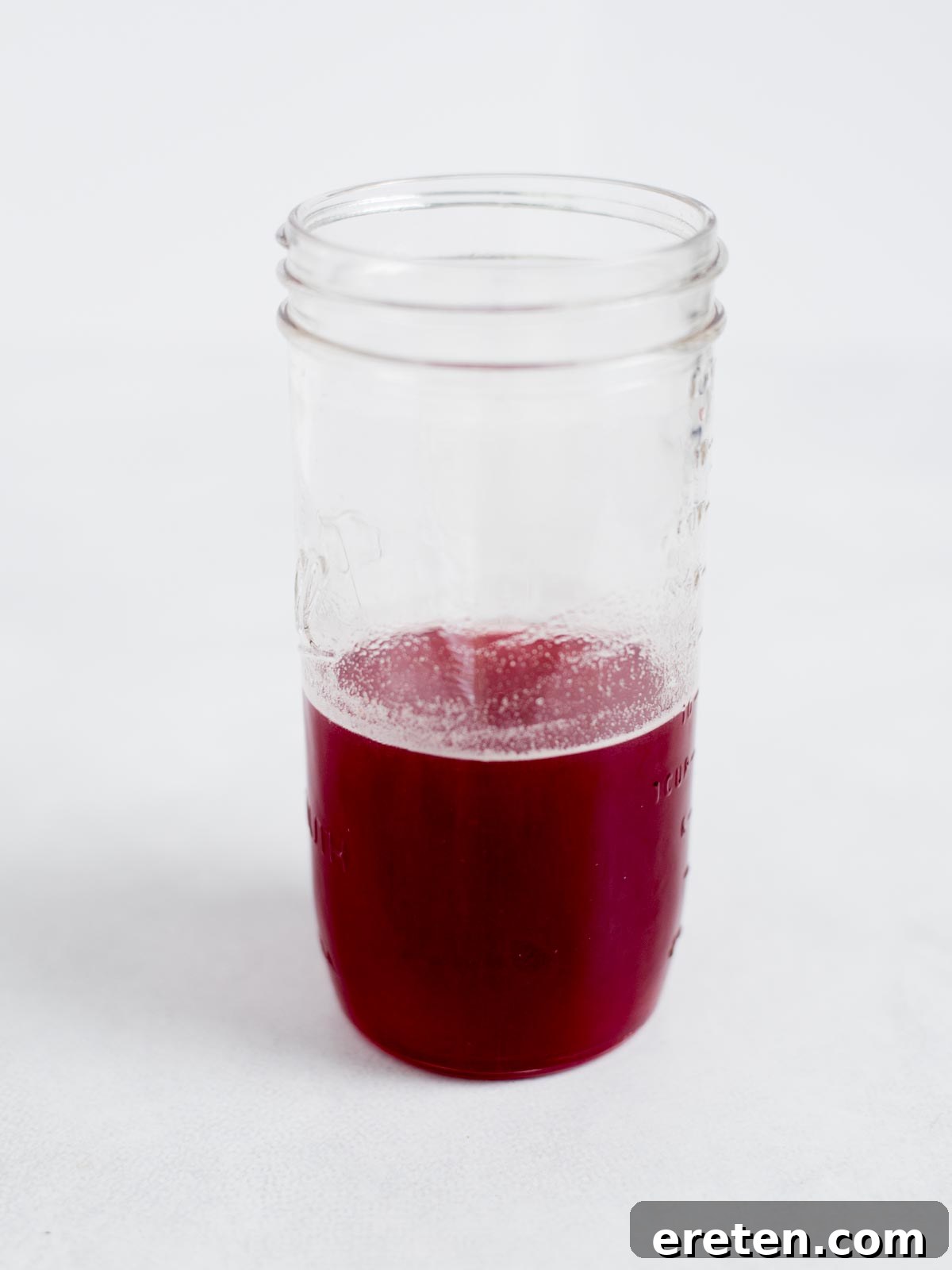 A small, clear glass jar containing a precious amount of bright, concentrated red raspberry syrup, freshly strained and ready for use. It sits neatly on a clean white surface with a simple, unfocused white background, emphasizing the syrup's vibrant color and clarity.