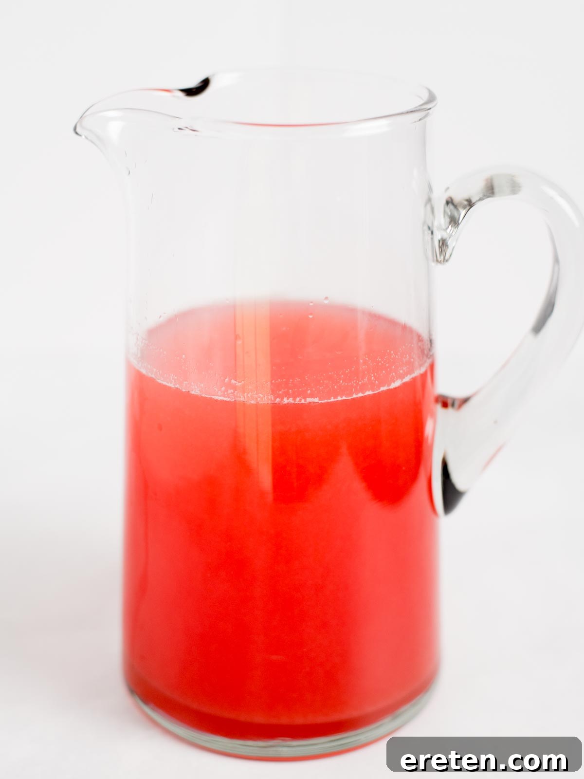 A gleaming clear glass pitcher, half-filled with the vibrant, inviting hue of bright red homemade raspberry lemonade. The pitcher stands proudly against a simple white background, its contents promising cool refreshment.