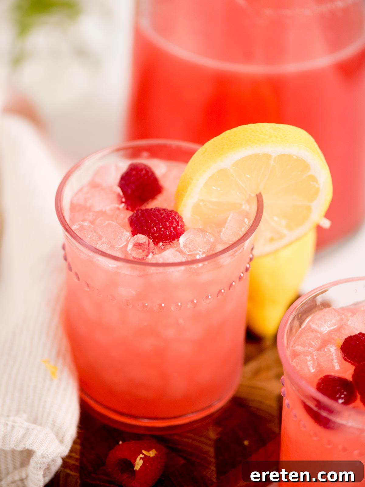 A tall, elegant glass of vibrant pink raspberry lemonade, generously filled with crushed ice and garnished with two fresh raspberries and a thin lemon slice. In the soft, blurred background, a matching pitcher of the same refreshing drink and a neatly folded white cloth complete the inviting scene, evoking a perfect summer moment.