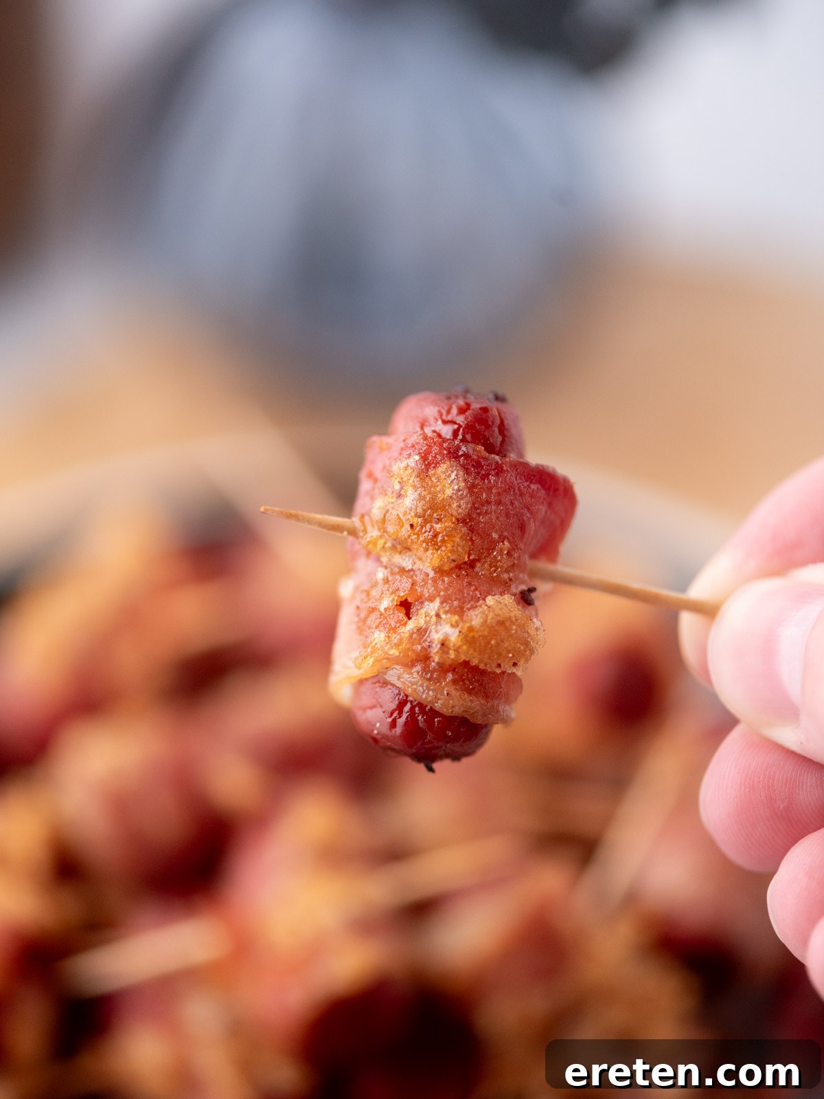 Golden Glazed Bacon Bites 2 A hand holds a toothpick skewering a small sausage wrapped in crispy bacon, with more bacon-wrapped sausages blurred in the background.