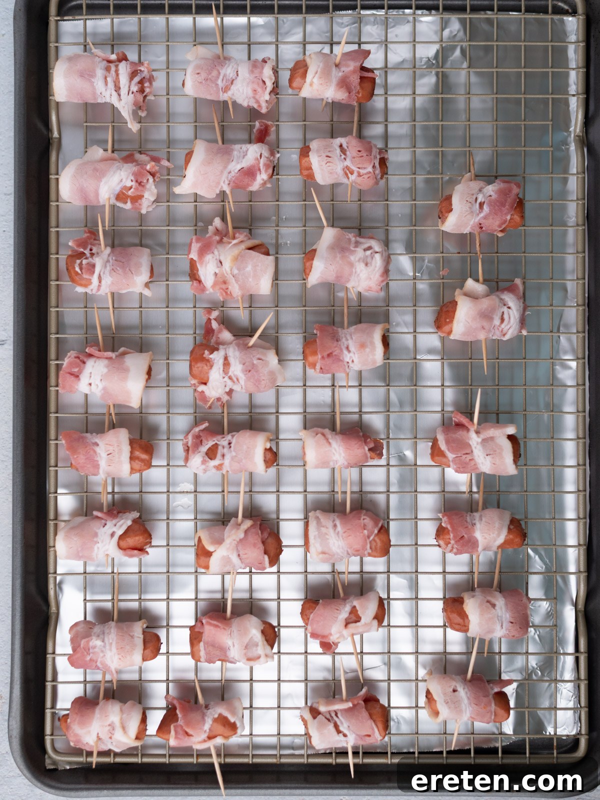 Golden Glazed Bacon Bites 4 Raw bacon-wrapped little smokies skewered with toothpicks are arranged in rows on a wire rack over a foil-lined baking sheet, ready to be cooked.