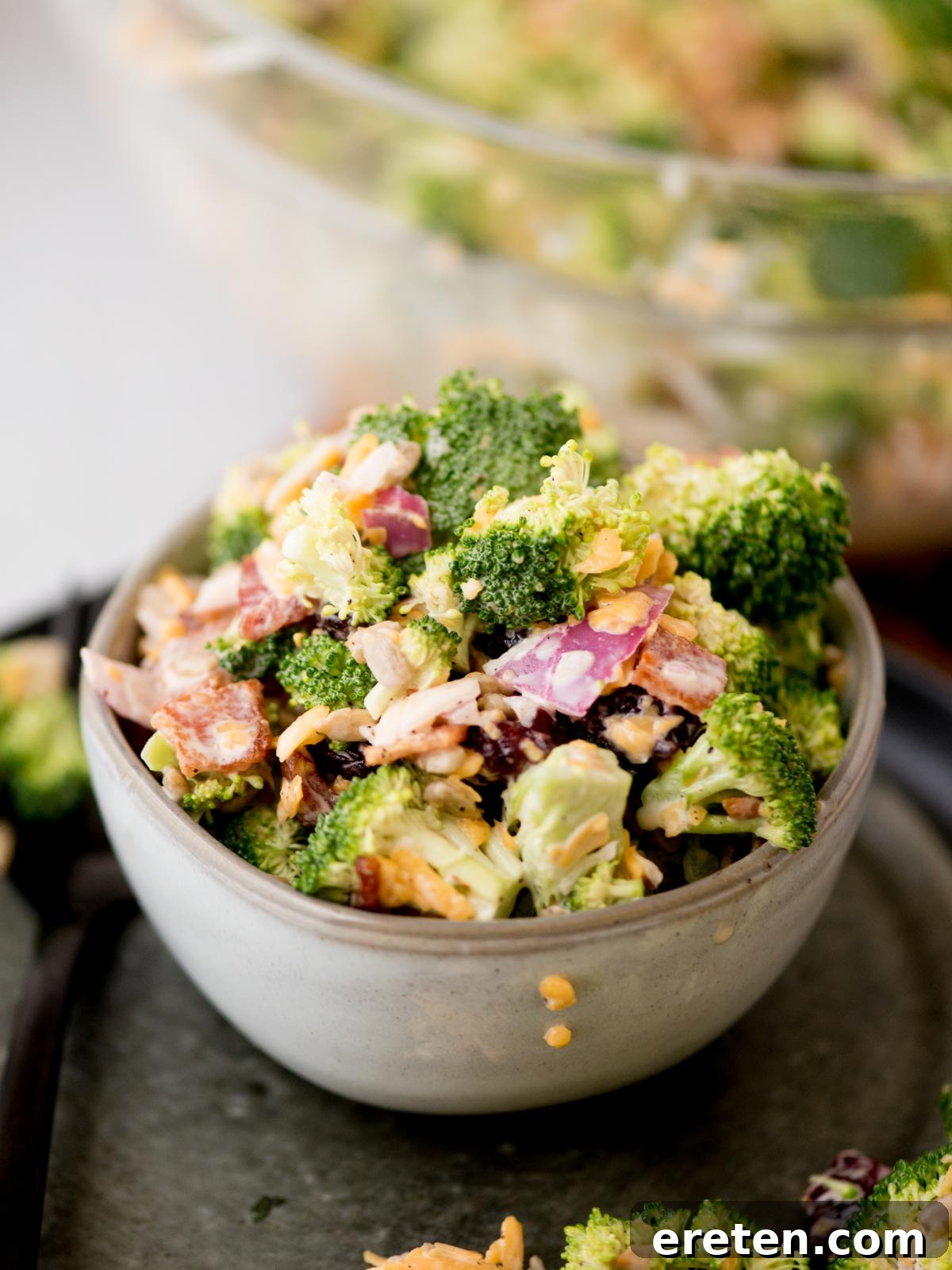 A vibrant bowl of broccoli salad, featuring crisp green broccoli florets, crumbled bacon, sharp shredded cheddar cheese, diced red onion, and sweet dried cranberries, all coated in a rich, creamy dressing. The bowl sits on a dark serving tray, with a larger, blurred bowl of salad in the background, suggesting a generous portion.