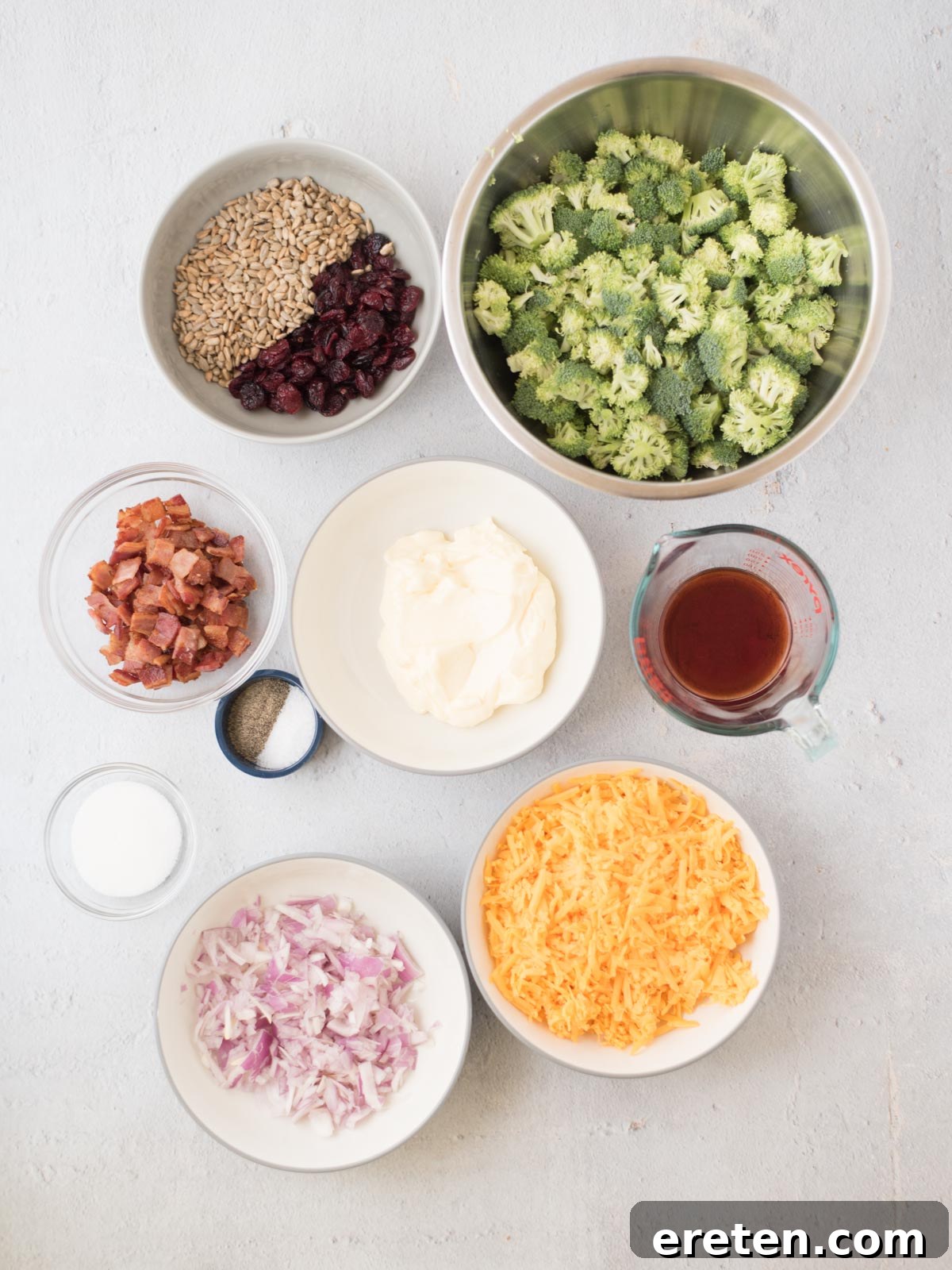 A selection of fresh ingredients laid out on a rustic wooden table, ready for a broccoli salad. The display includes vibrant green chopped broccoli florets, golden shredded sharp cheddar cheese, creamy mayonnaise in a bowl, finely diced red onion, crispy cooked bacon bits, a small pile of roasted sunflower seeds, a handful of ruby-red dried cranberries, a small bowl of granulated sugar, and a measuring cup containing red wine vinegar.