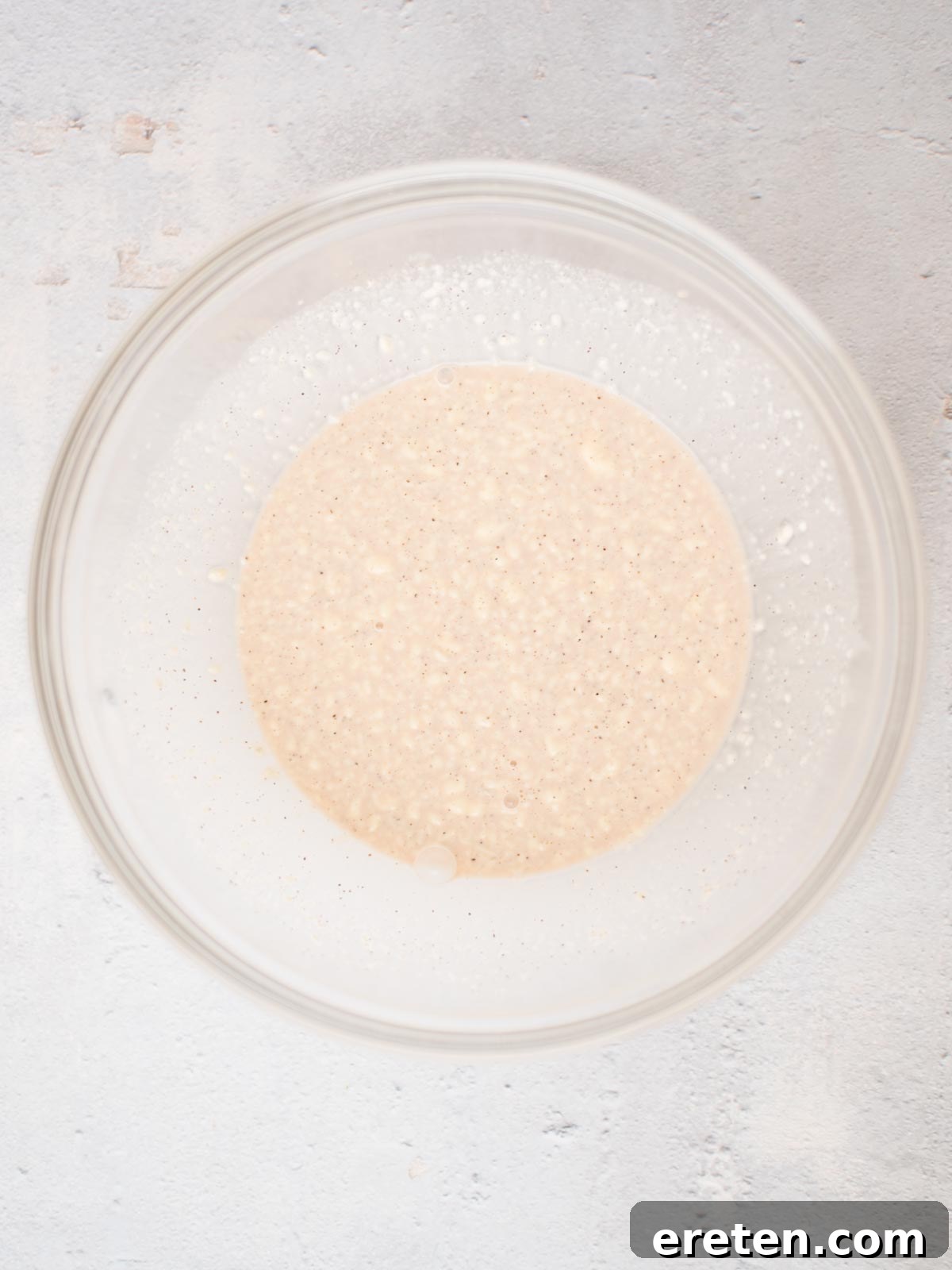 A close-up shot of a clear glass mixing bowl filled with a creamy, pale yellow dressing mixture. The surface shows small whisk marks and air bubbles, indicating it has just been freshly prepared. The bowl is resting on a light-colored kitchen counter.