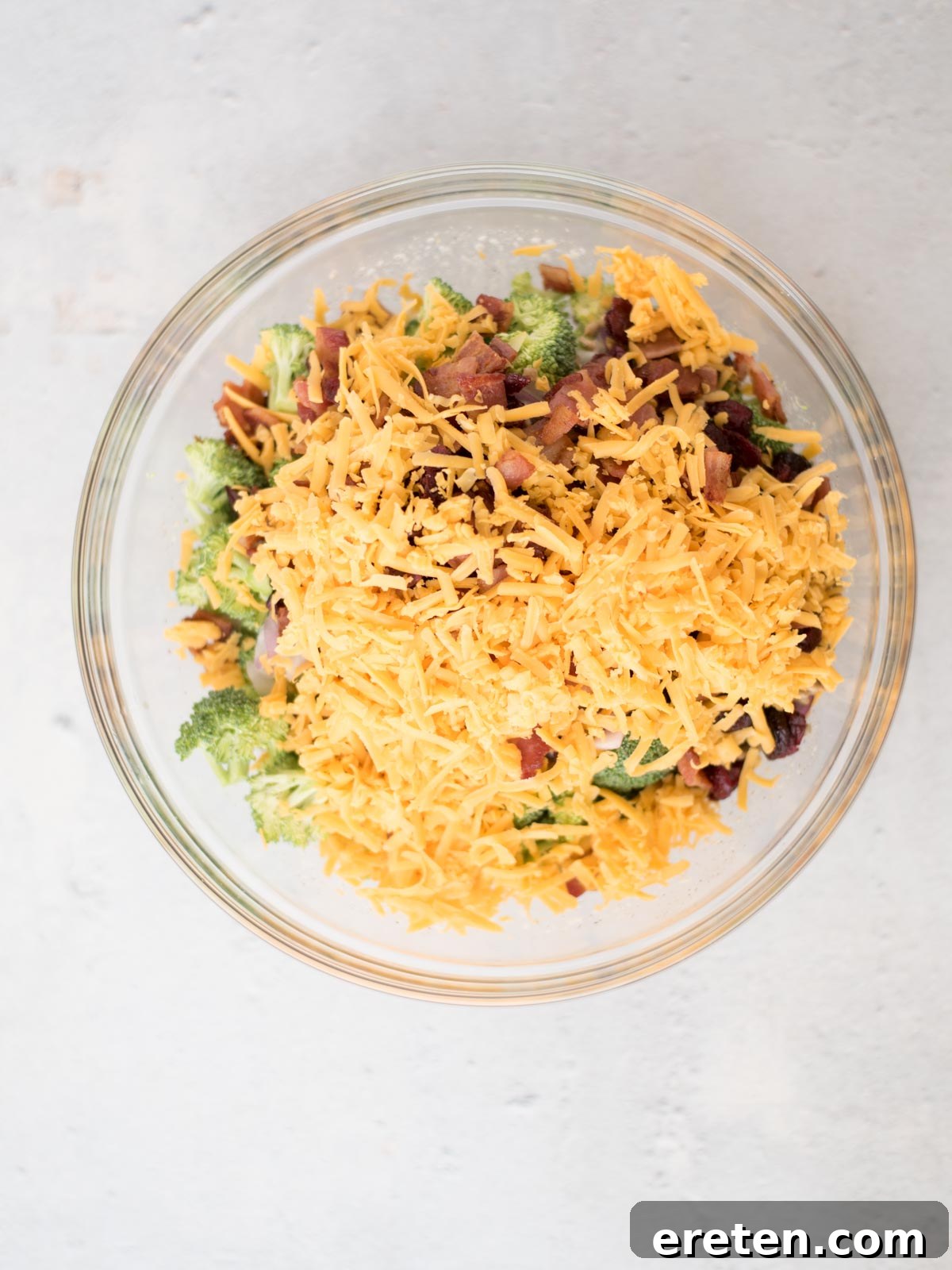 A clear glass mixing bowl filled with a vibrant, freshly prepared broccoli salad. The salad features finely chopped green broccoli, crispy bacon bits, bright red dried cranberries, and generous amounts of shredded cheddar cheese, all thoroughly mixed and coated in a creamy dressing. The bowl is set against a softly lit, light background.
