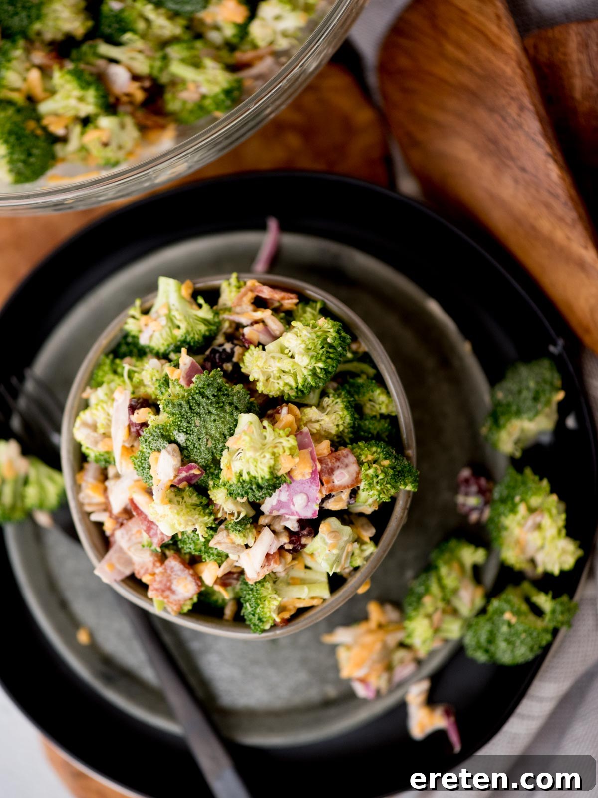 A close-up shot of a small, elegant grey bowl filled with a delightful broccoli salad. The salad is rich with chopped green broccoli, vibrant red onion pieces, crispy bacon bits, and generous amounts of shredded cheese. A fork rests delicately beside the bowl on a dark, textured plate, with a rustic wooden board subtly visible in the soft-focus background.