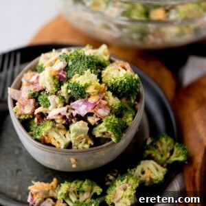 A bowl of broccoli salad with chopped red onions, shredded cheese, and bits of bacon. The salad is in a small grey bowl on a dark plate, with some salad spilled around it. A larger bowl with more salad is blurred in the background.