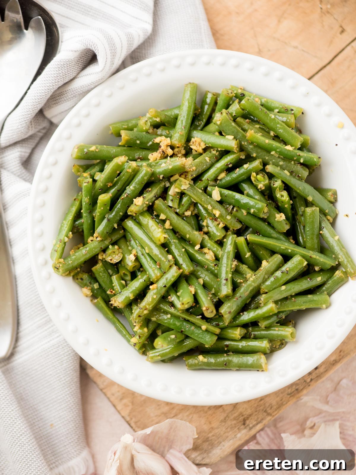 A white bowl filled with perfectly cooked green beans, glistening with minced garlic and seasonings, resting on a rustic wooden surface next to a striped linen towel, a sleek metal serving spoon, and fresh garlic cloves.