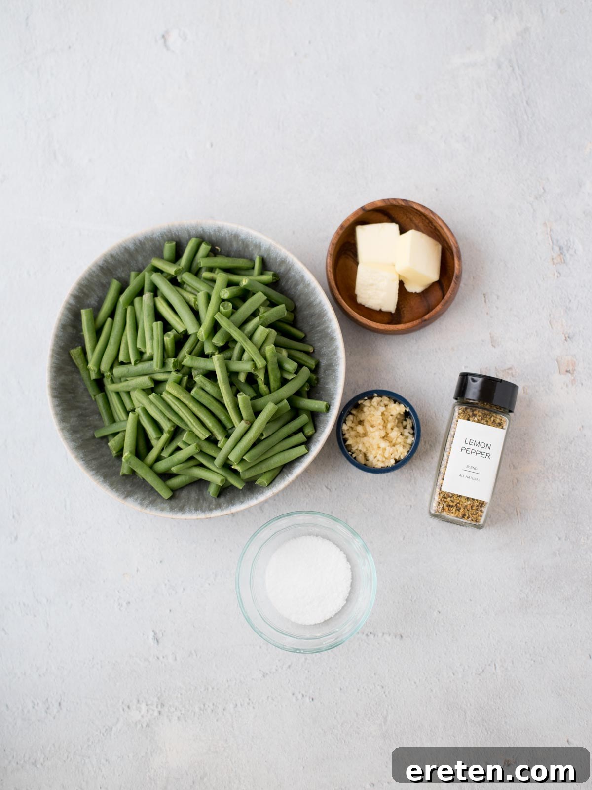 A beautifully arranged still life of ingredients: a bowl filled with neatly cut fresh green beans, a small dish holding cubes of golden butter, a tiny bowl brimming with freshly minced garlic, a distinct jar of lemon pepper seasoning, and a small pinch bowl of coarse salt, all laid out on a clean, light-colored surface.