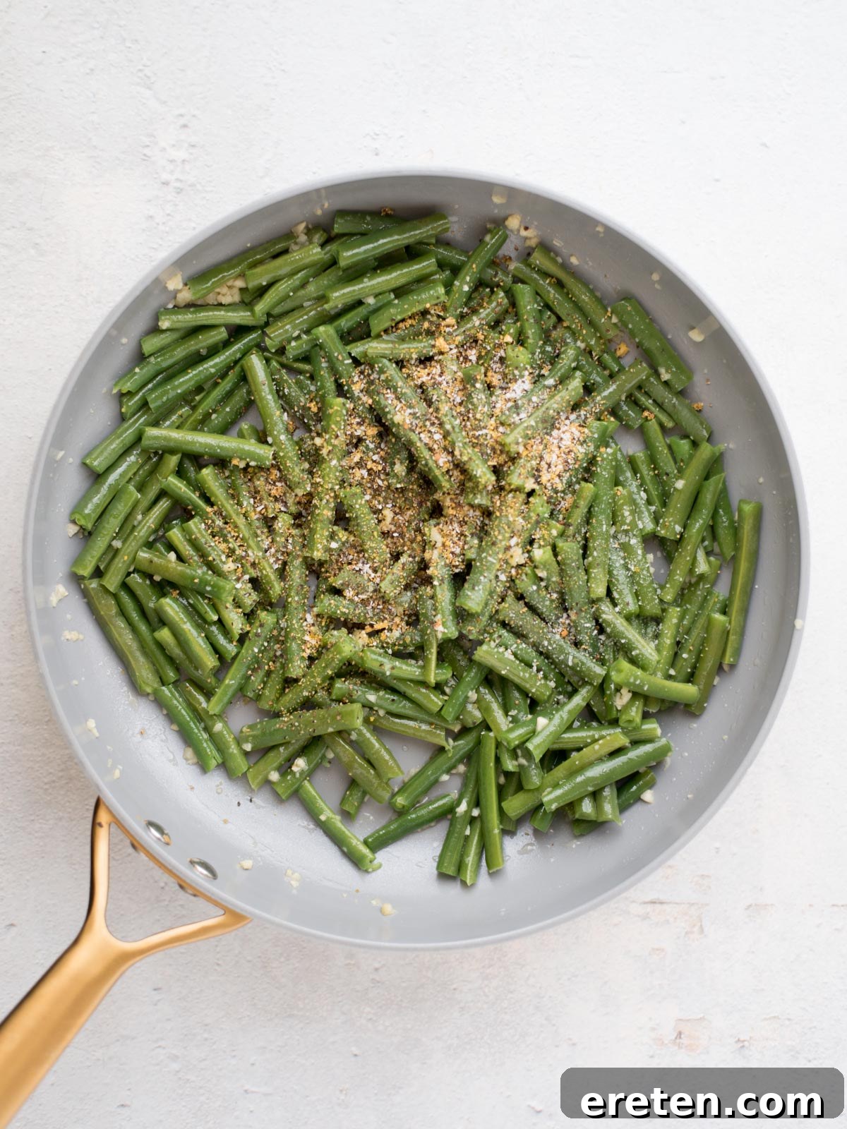 A vibrant close-up of a frying pan filled with beautifully cooked green beans, expertly cut into pieces, generously topped with a sprinkle of aromatic seasoning and golden breadcrumbs (if desired, not explicit in the text but common visually), all resting on a clean, light-colored kitchen surface.