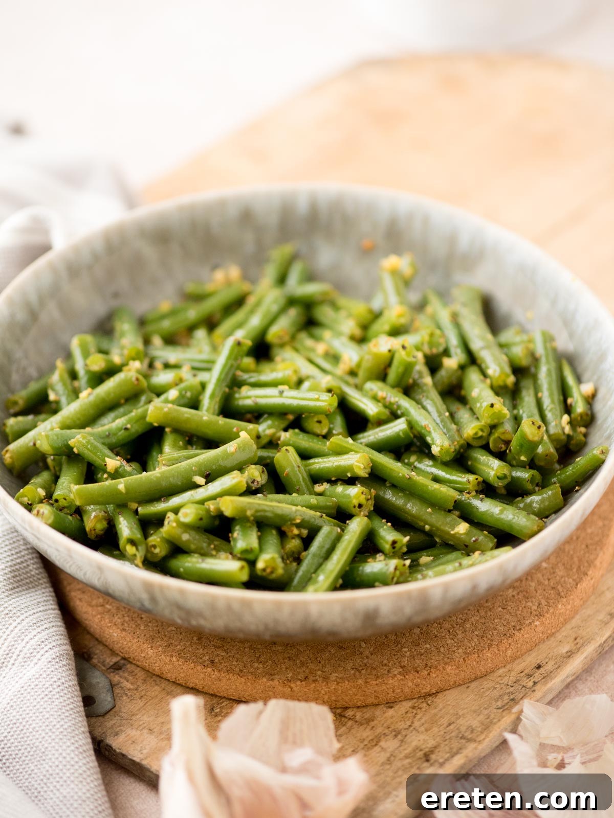 A charming ceramic bowl generously filled with perfectly cooked green beans, richly tossed with fragrant garlic and a medley of aromatic spices. The bowl is positioned invitingly on a warm, wooden surface, with a scattering of fresh garlic cloves nearby to emphasize the key flavor.