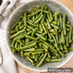 A bowl of cooked green beans cut into pieces, seasoned with herbs and garlic, sits on a wooden surface next to a striped cloth.