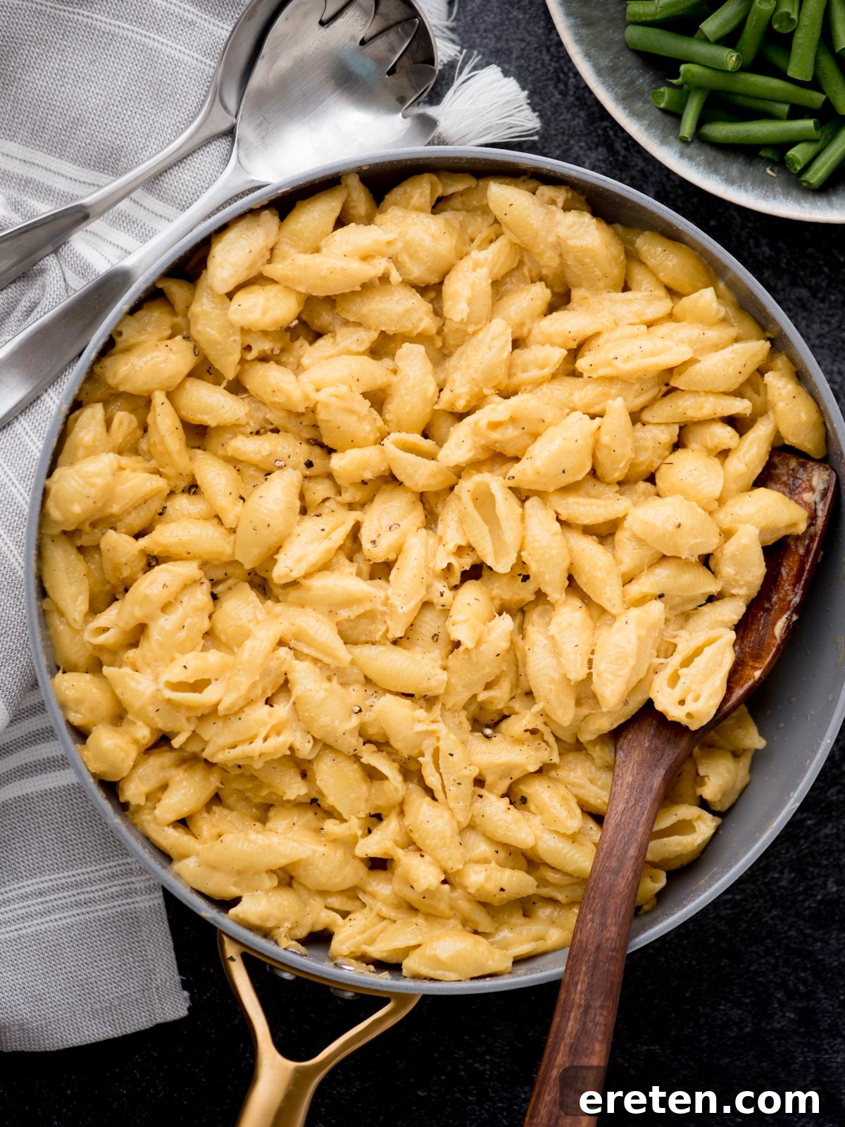 A skillet filled with creamy macaroni and cheese made from shell pasta, with a wooden spoon resting in it. A pair of serving utensils and a plate of green beans are beside the skillet on a dark surface, highlighting the golden hue of the dish.