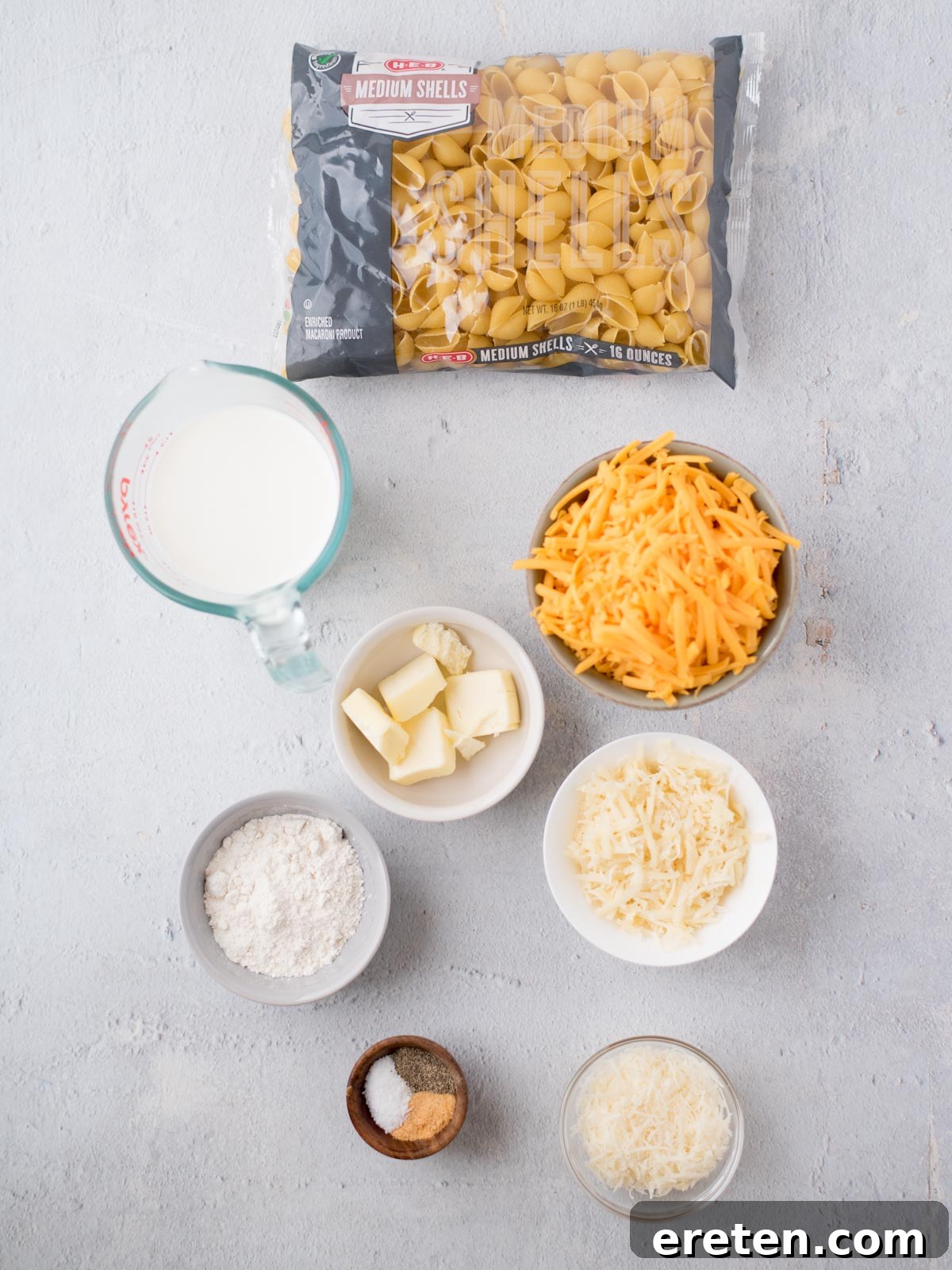 Top view of ingredients for macaroni and cheese on a light surface. Visible items include a bag of medium shell pasta, shredded cheddar cheese, grated Parmesan cheese, butter, flour, a measuring cup of milk, and small bowls of salt and nutmeg, all perfectly arranged.
