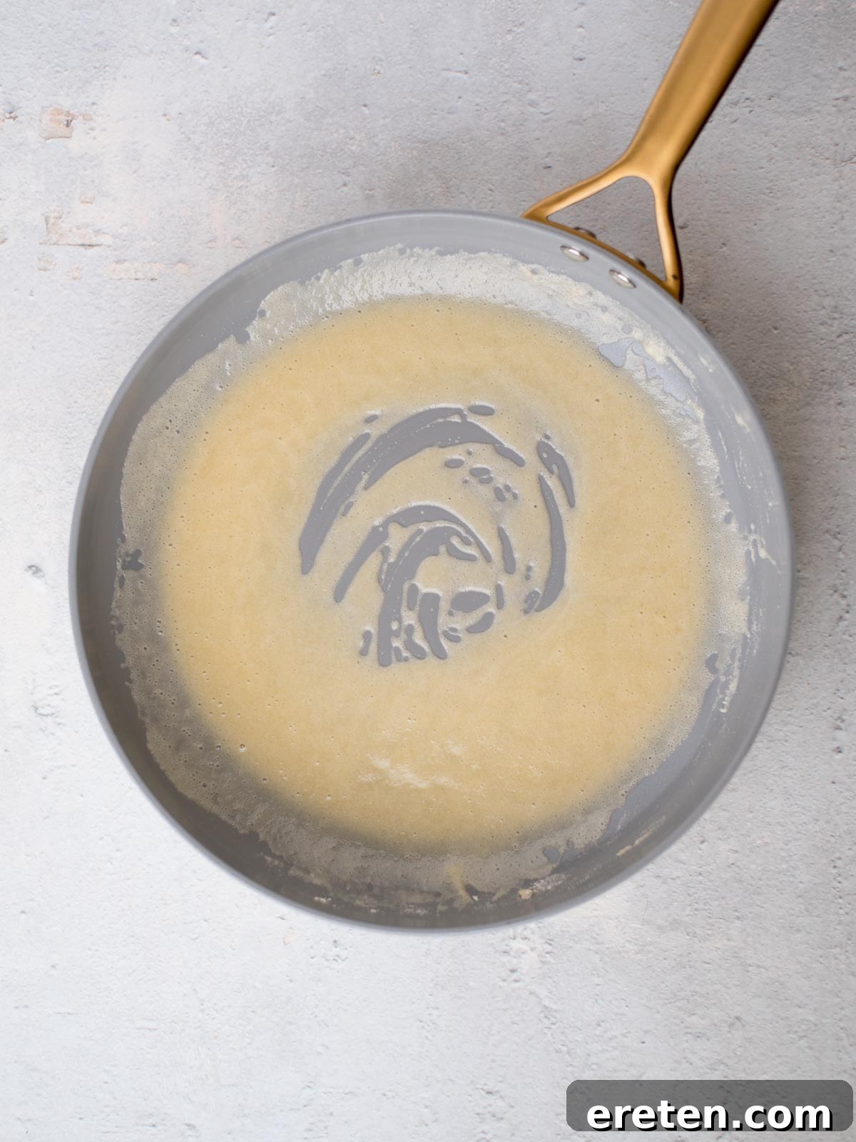 A non-stick frying pan with a swirl of beige batter or sauce on a light gray, textured surface. The handle is metallic and golden-colored, showcasing the smoothly cooked roux.