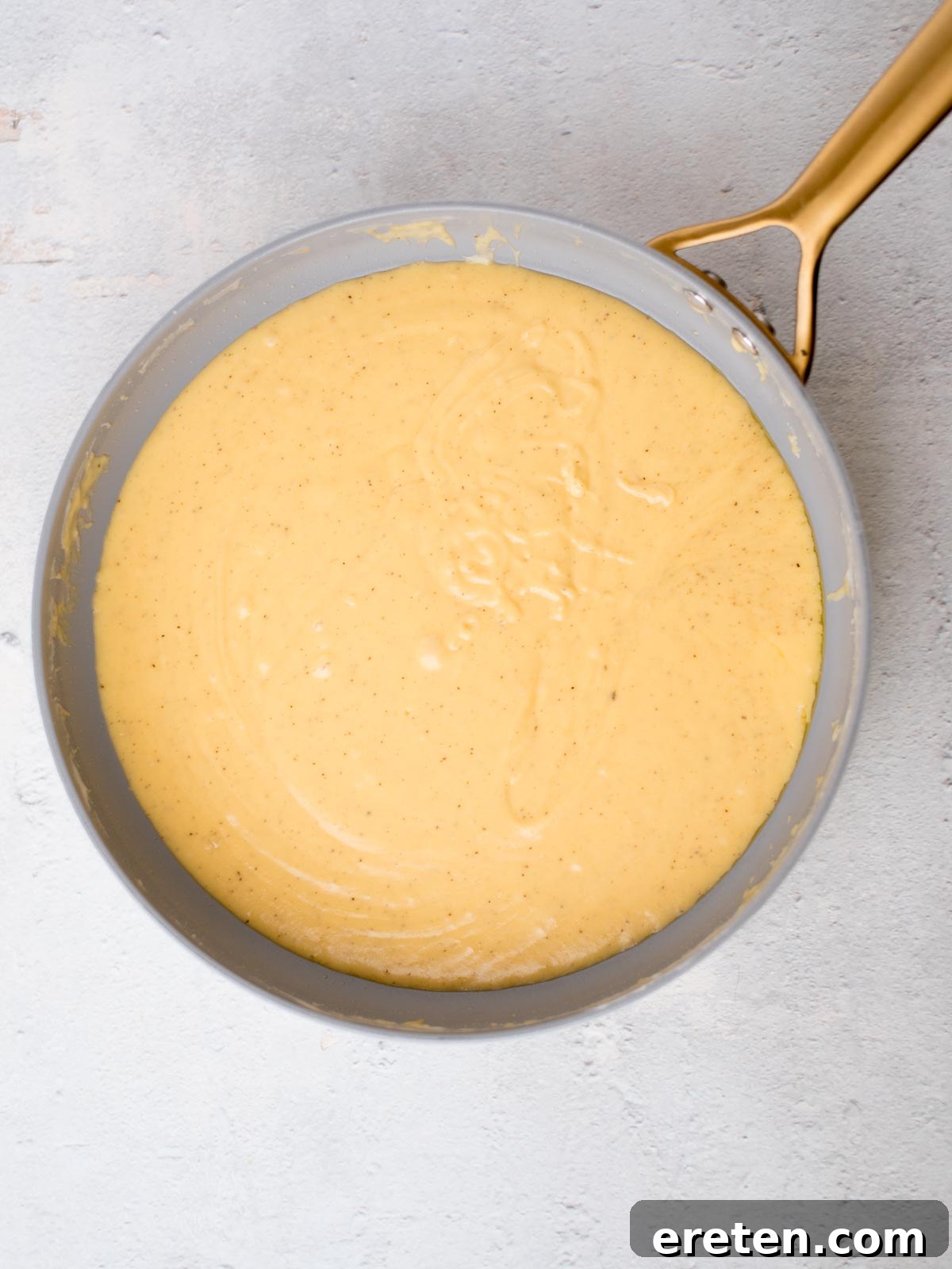 A round pan filled with a creamy, yellowish batter sits on a light gray surface. The batter appears smooth and slightly thick, with visible specks, possibly indicating spices or seasoning, highlighting the rich cheese sauce.